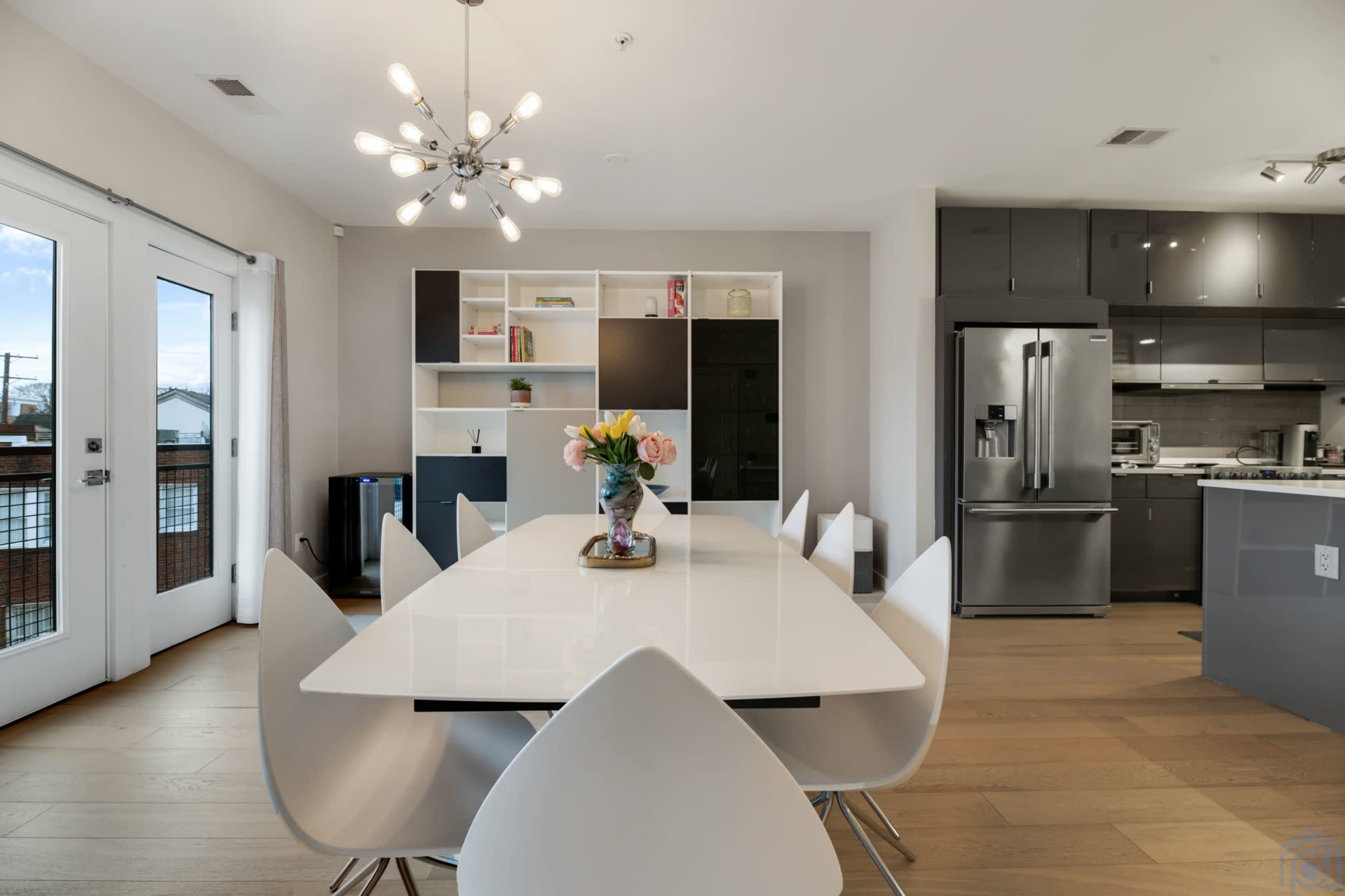 A modern dining area features a large white table surrounded by white chairs, with a bouquet of flowers at the center and an open kitchen visible in the background.