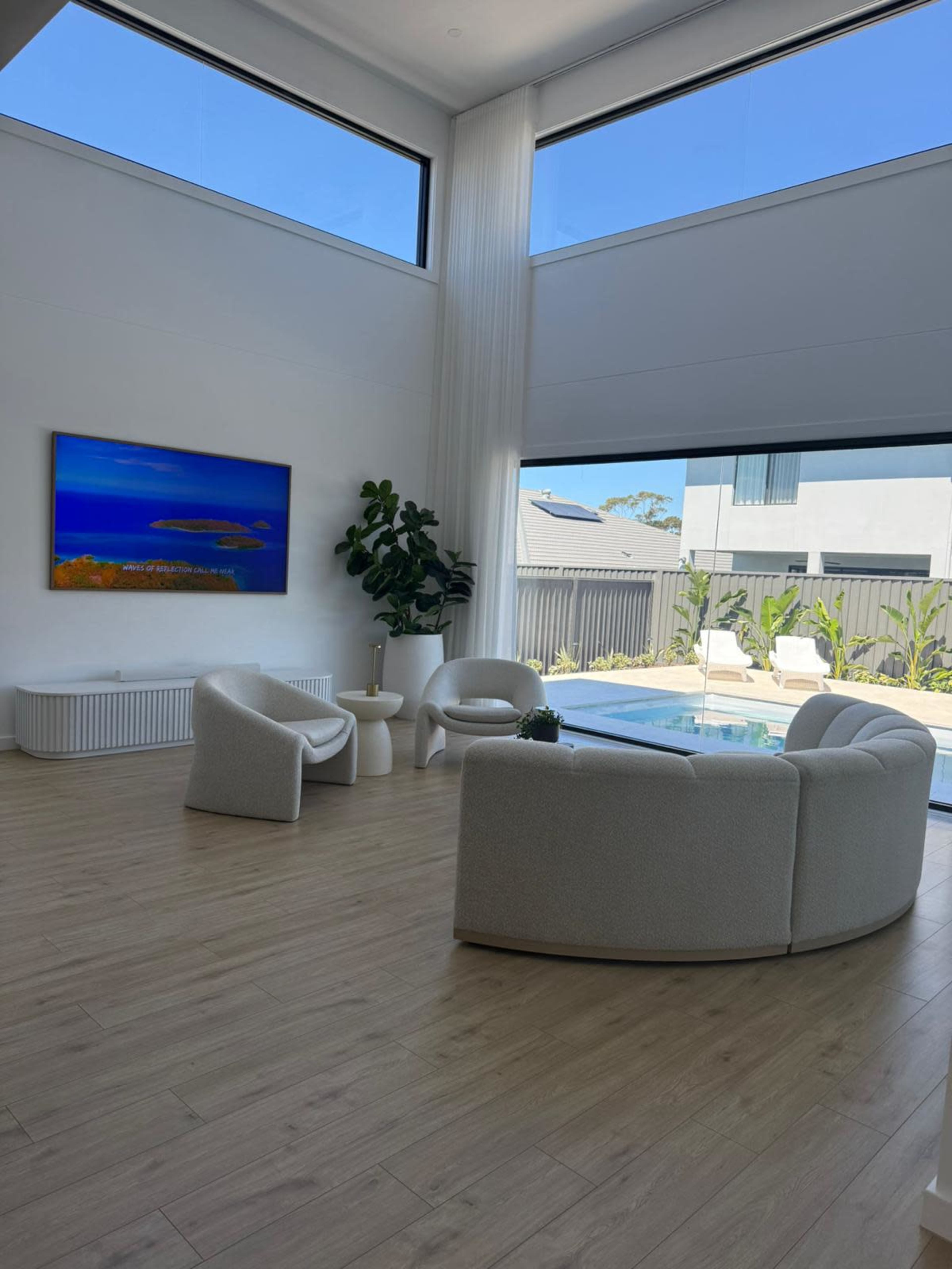 The image shows a modern living room with two white, curved sofas facing a glass wall that overlooks a pool and garden area, with a colorful painting on the wall.
