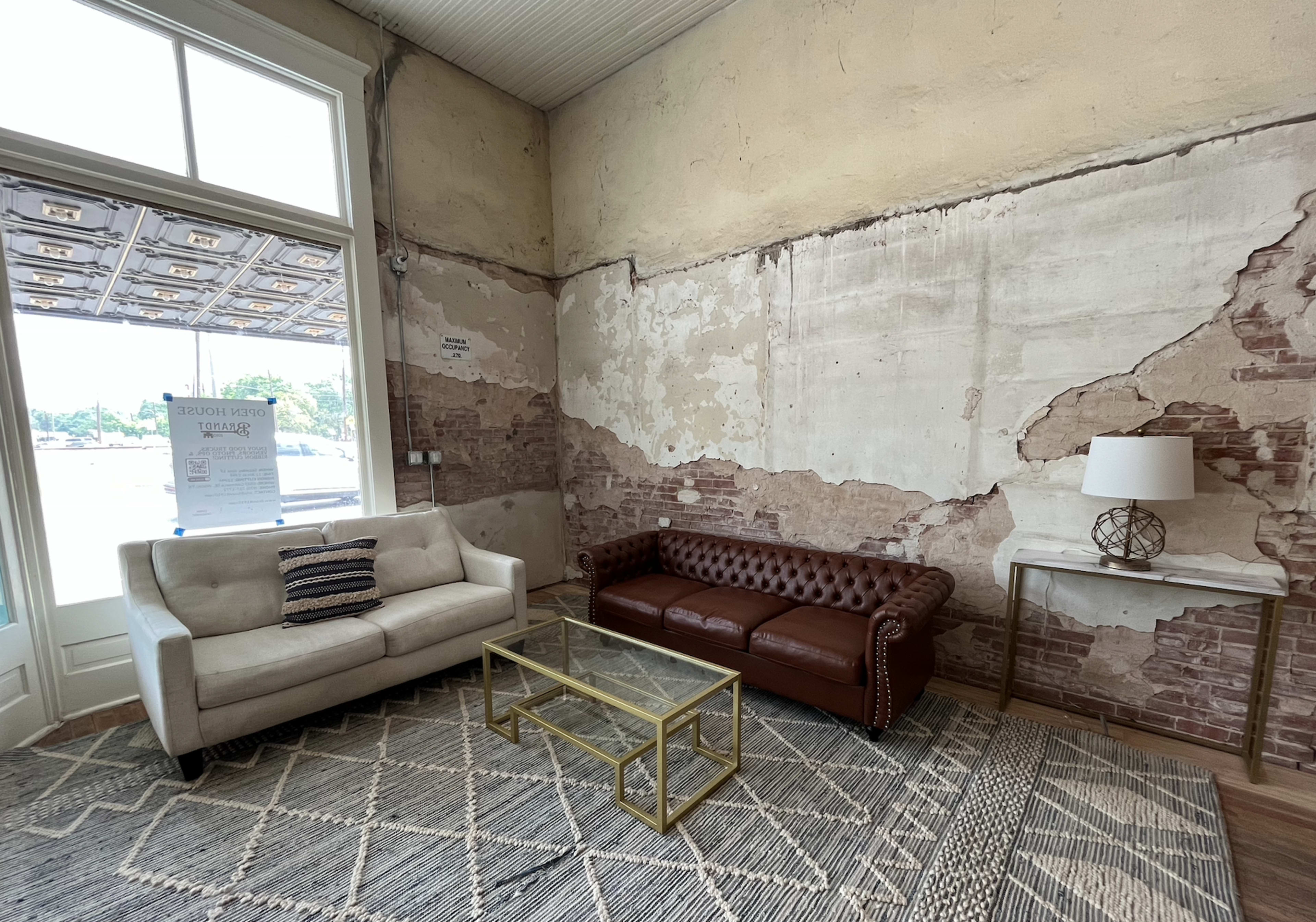 The image shows a living room with two sofas, one white and one brown leather, a glass coffee table, and a lamp on a side table, against a wall with exposed brick and peeling paint.