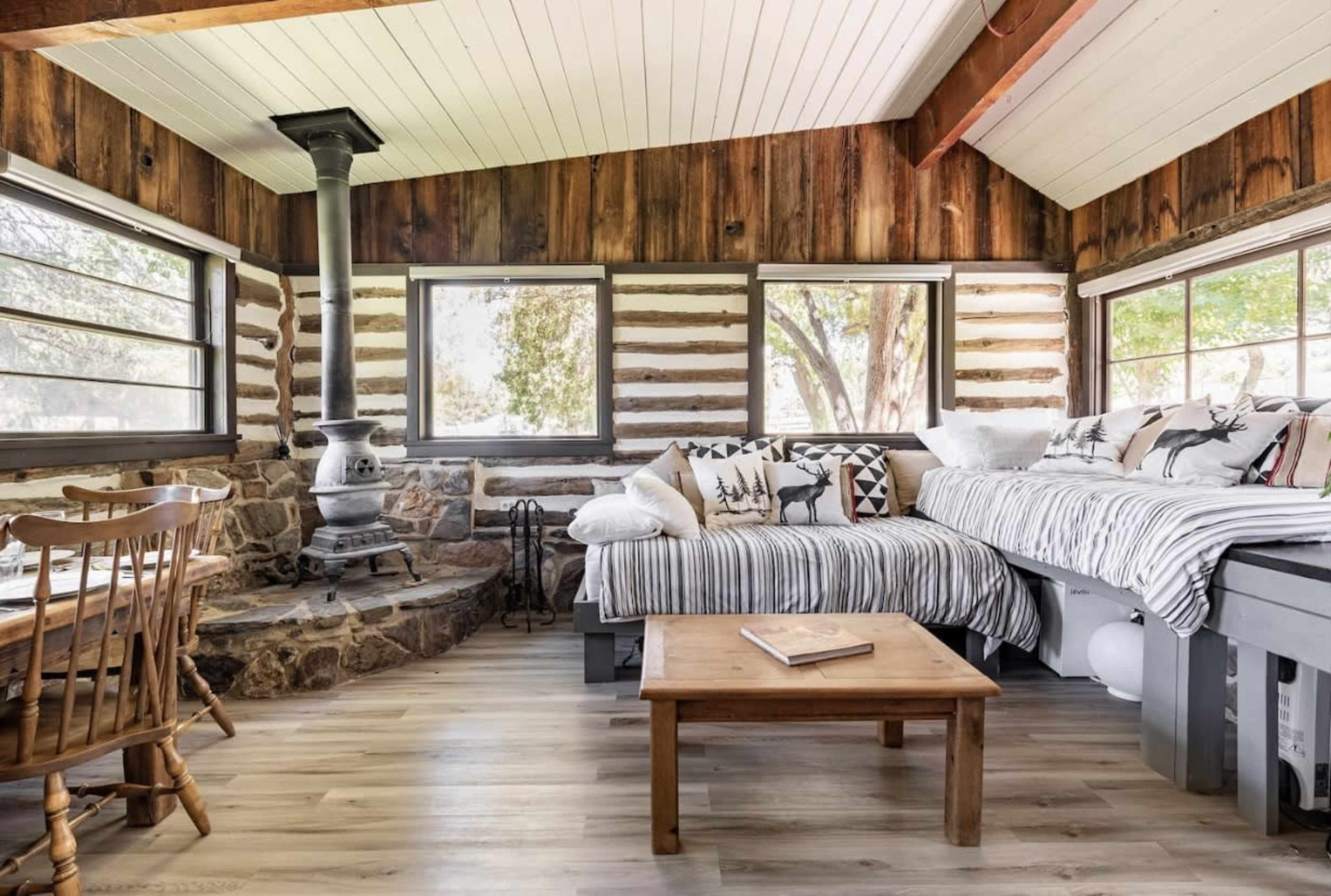 A cozy cabin interior featuring a wood stove, wooden walls, a coffee table, and a seating area with patterned pillows.