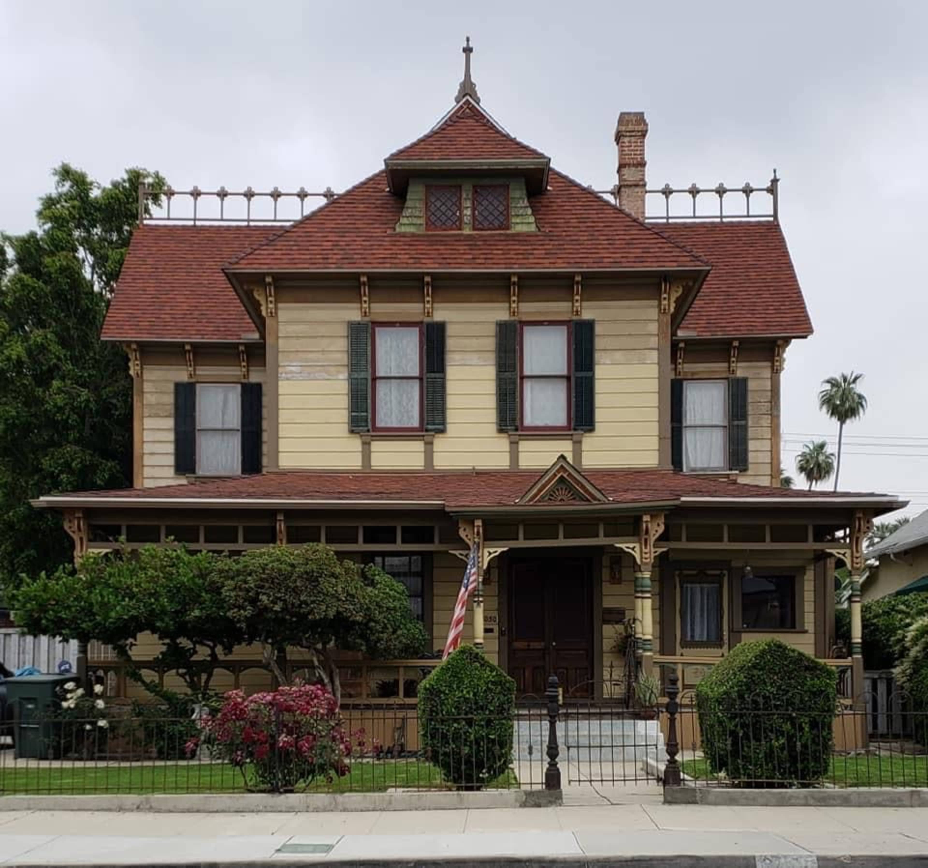 A Victorian-style house with a multi-gabled roof, ornate detailing, and a front porch surrounded by manicured bushes and a railing.