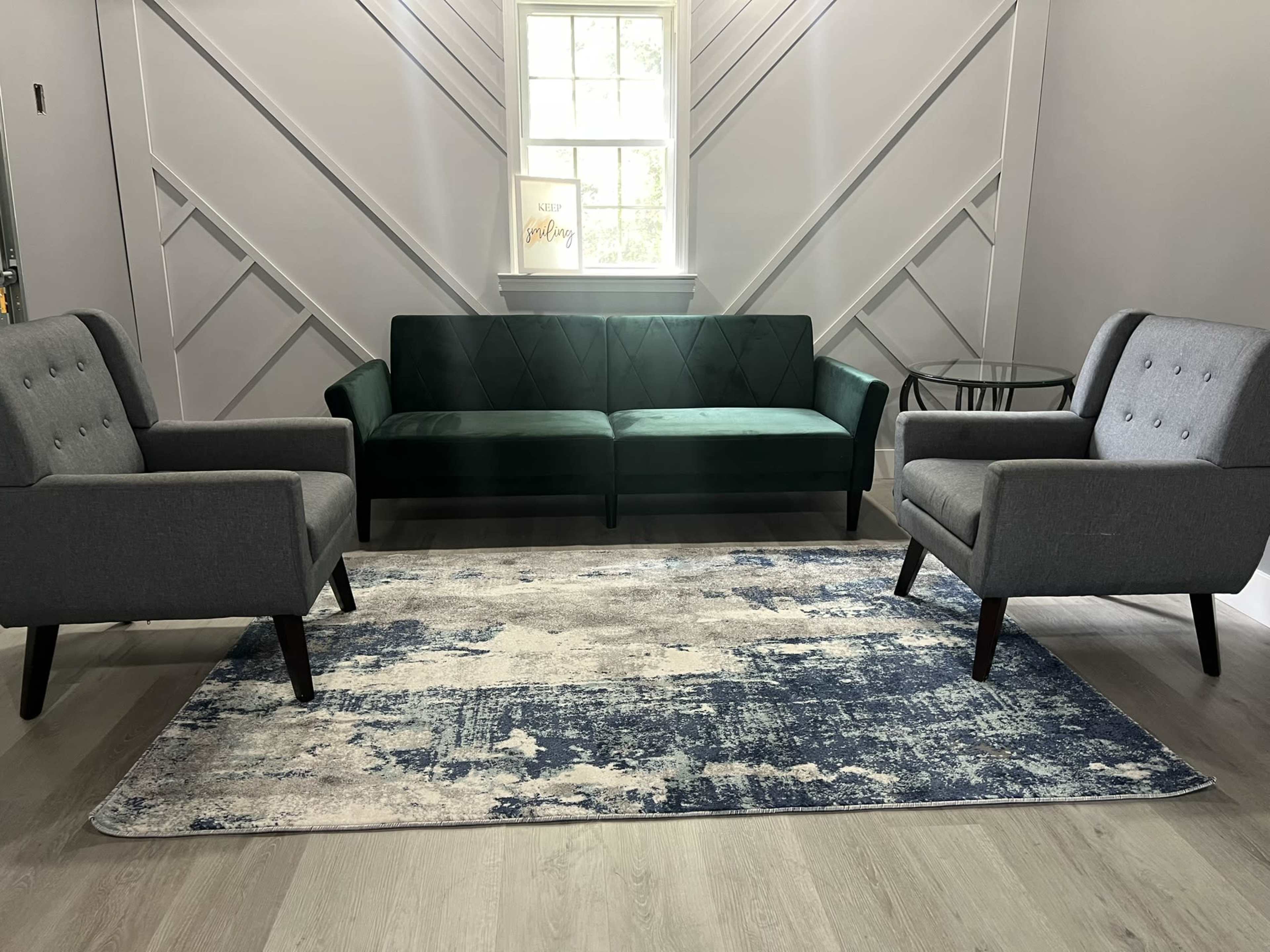 The image features a modern living room setup with a green couch, two grey armchairs, a small glass side table, and a patterned rug on a light wooden floor.