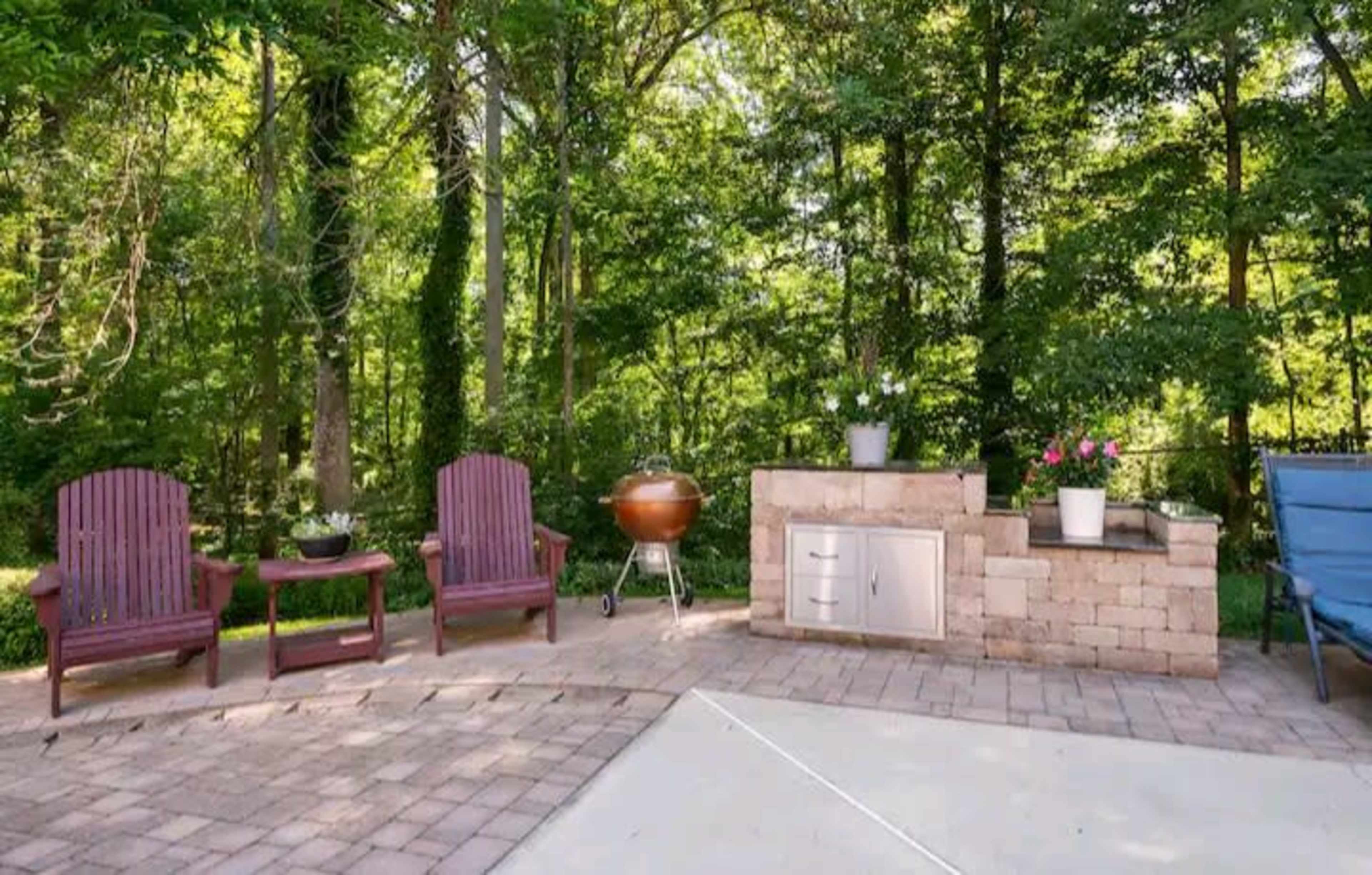 A patio area with two wooden chairs, a round grill, and a stone structure containing storage and potted plants, all surrounded by trees.