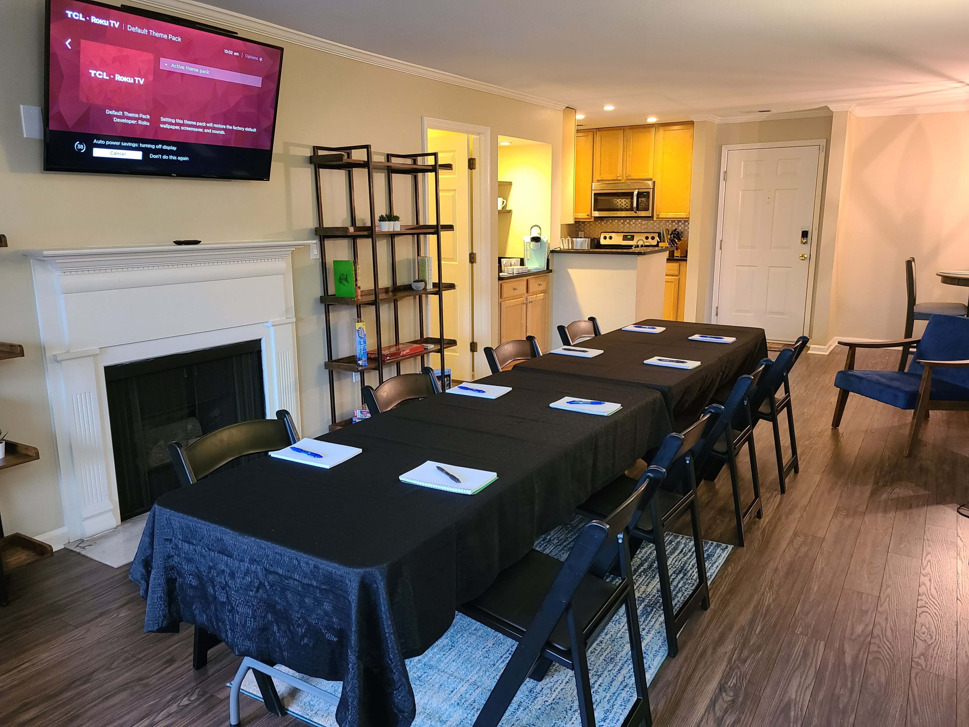 A long table with black cloth is set up in a meeting room, surrounded by black chairs, with a television and kitchen area visible in the background.
