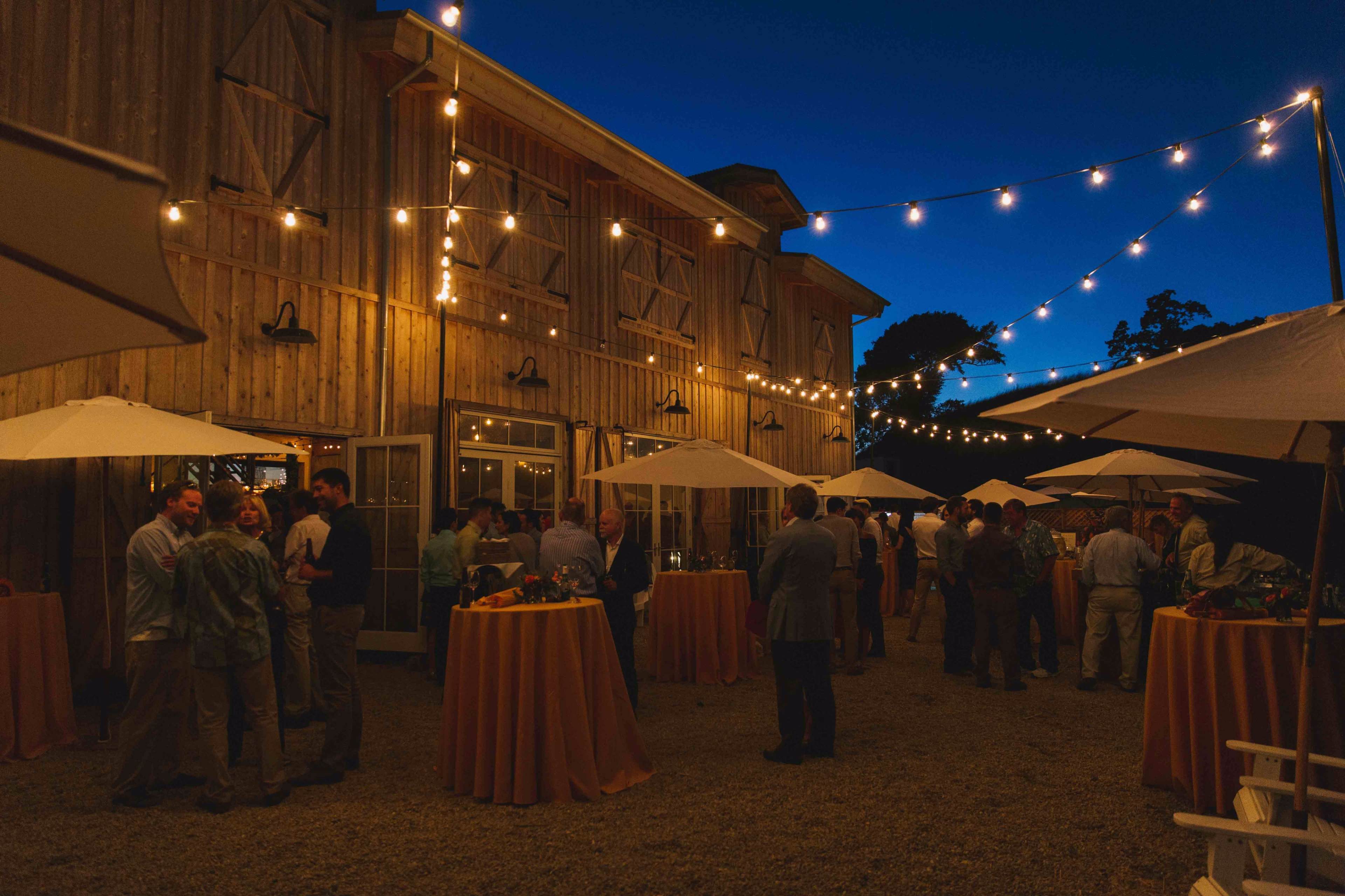 A gathering of guests is taking place outdoors near a barn, illuminated by string lights and accompanied by tables with umbrellas.