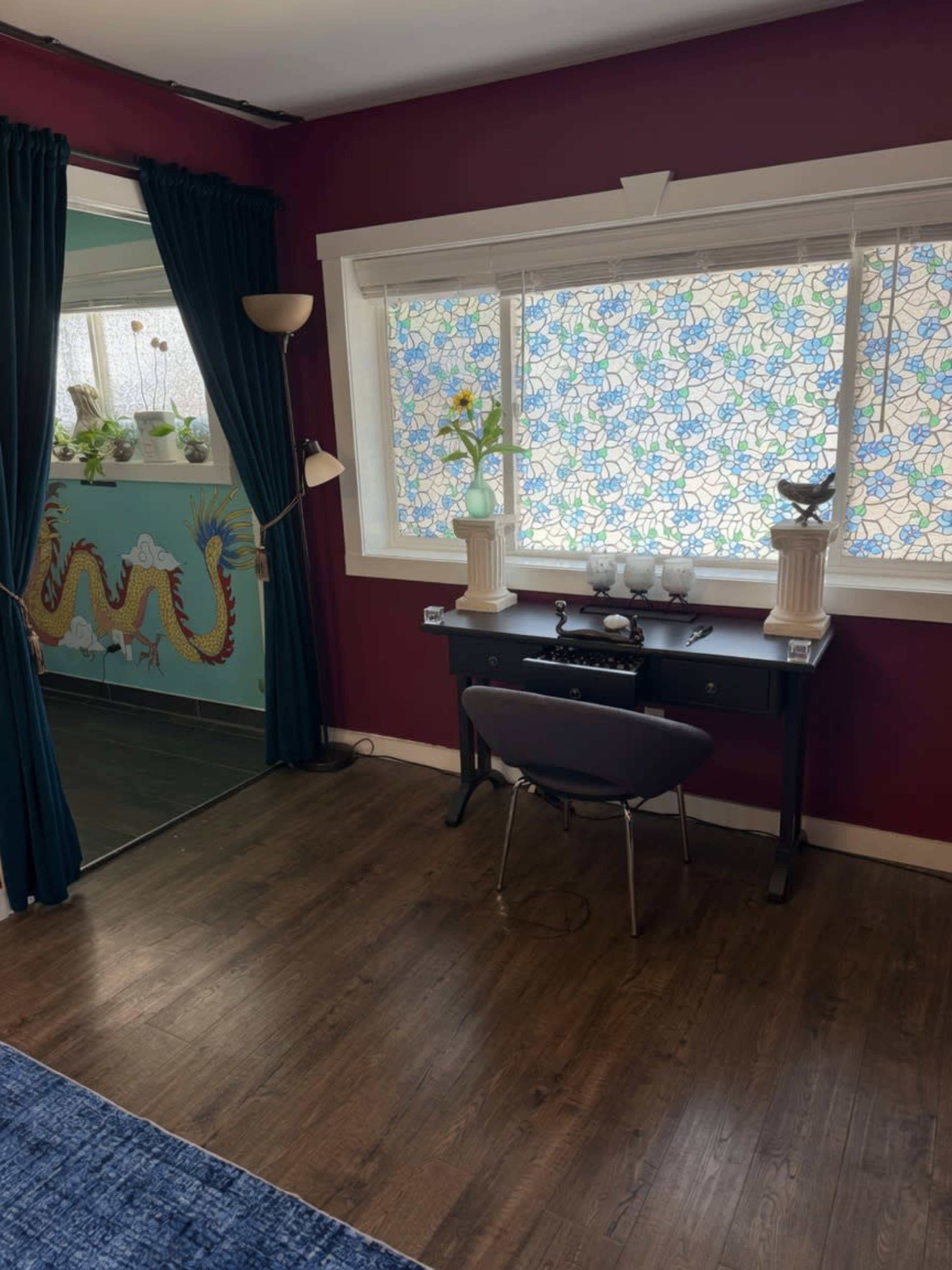 The image shows a room with a dark burgundy wall, a desk with decorative items, a chair, and a window adorned with colorful stained glass curtains, along with a mirror reflecting part of the room.