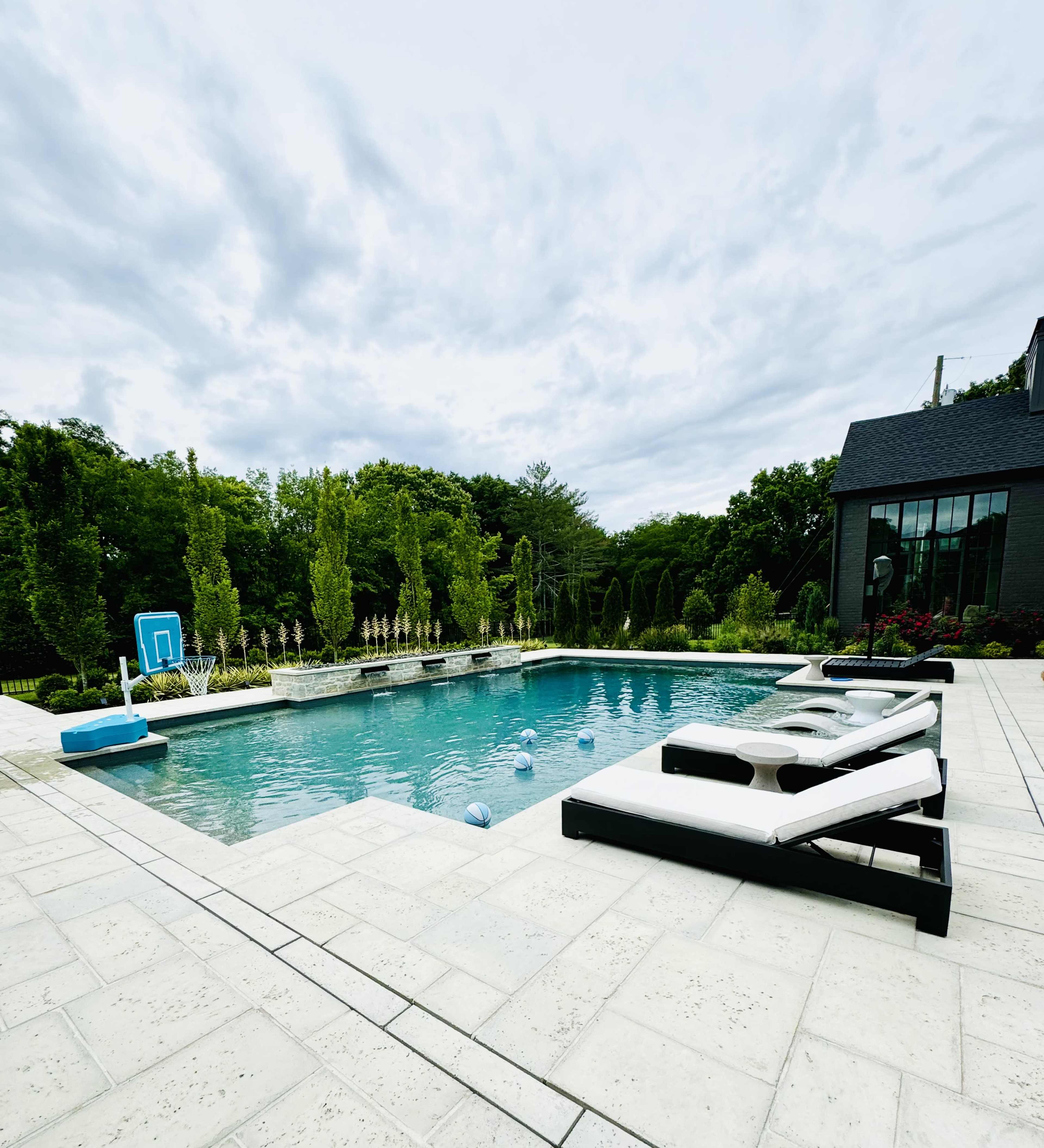 A rectangular swimming pool surrounded by a patio, with lounge chairs on one side and a basketball hoop on the opposite end.