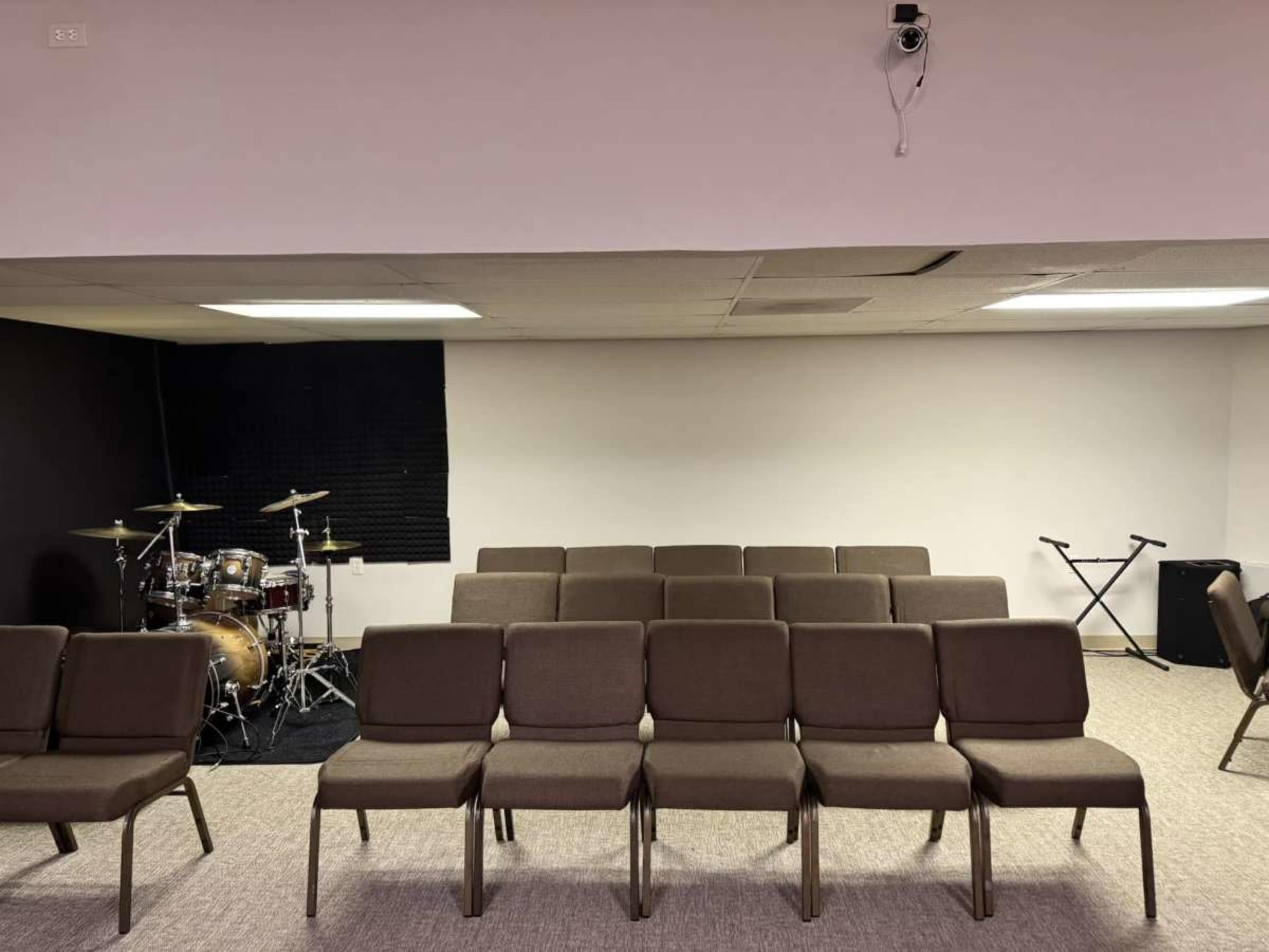 The image shows a room with several rows of chairs arranged facing a wall, a drum set in the corner, and a keyboard stand nearby.