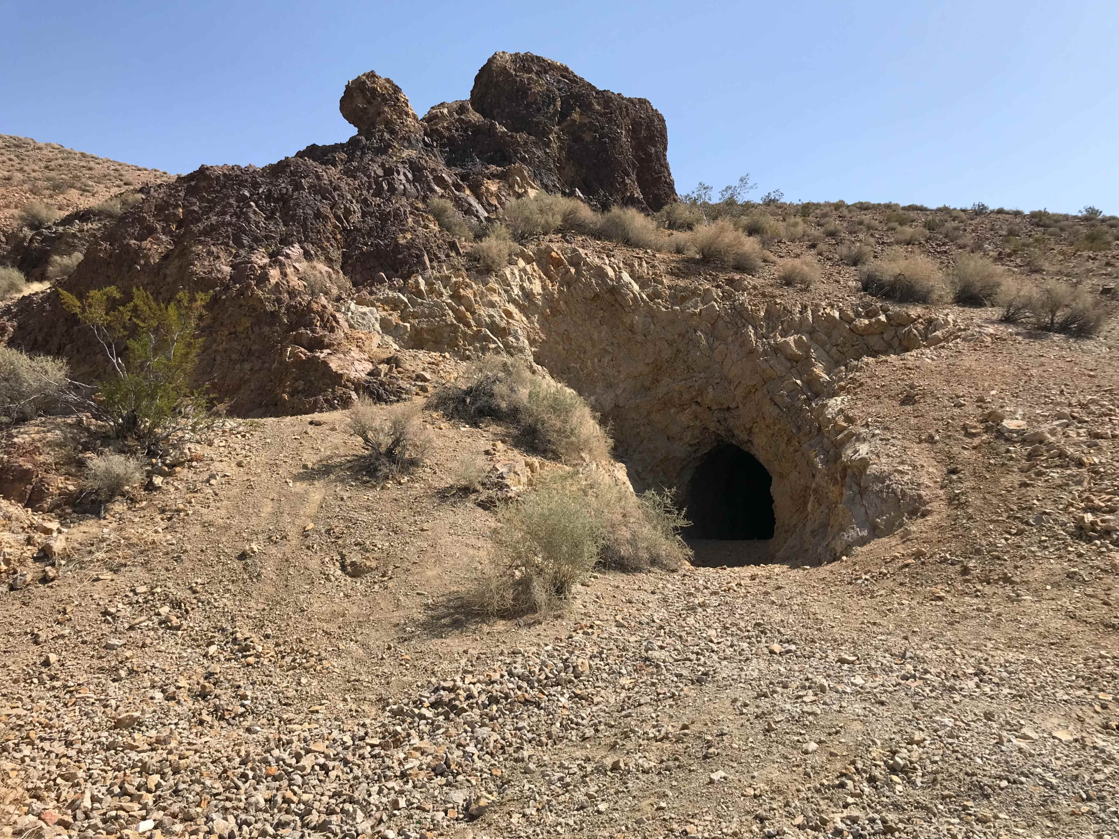 A rocky hillside with sparse vegetation features an entrance to a dark cave.