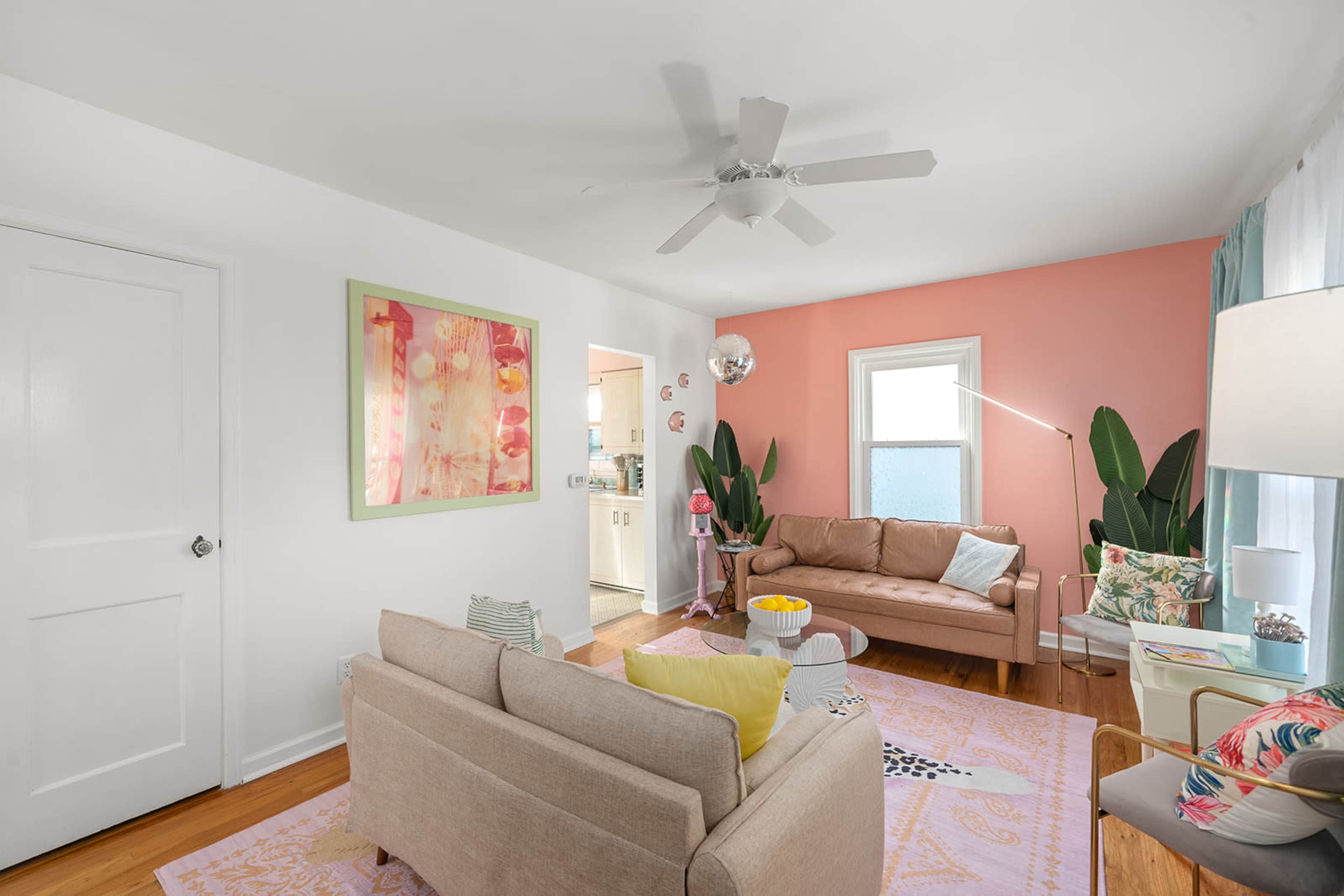 The living room features a beige sofa, a pink accent wall, and large green plants, with a glimpse of a bathroom in the background.