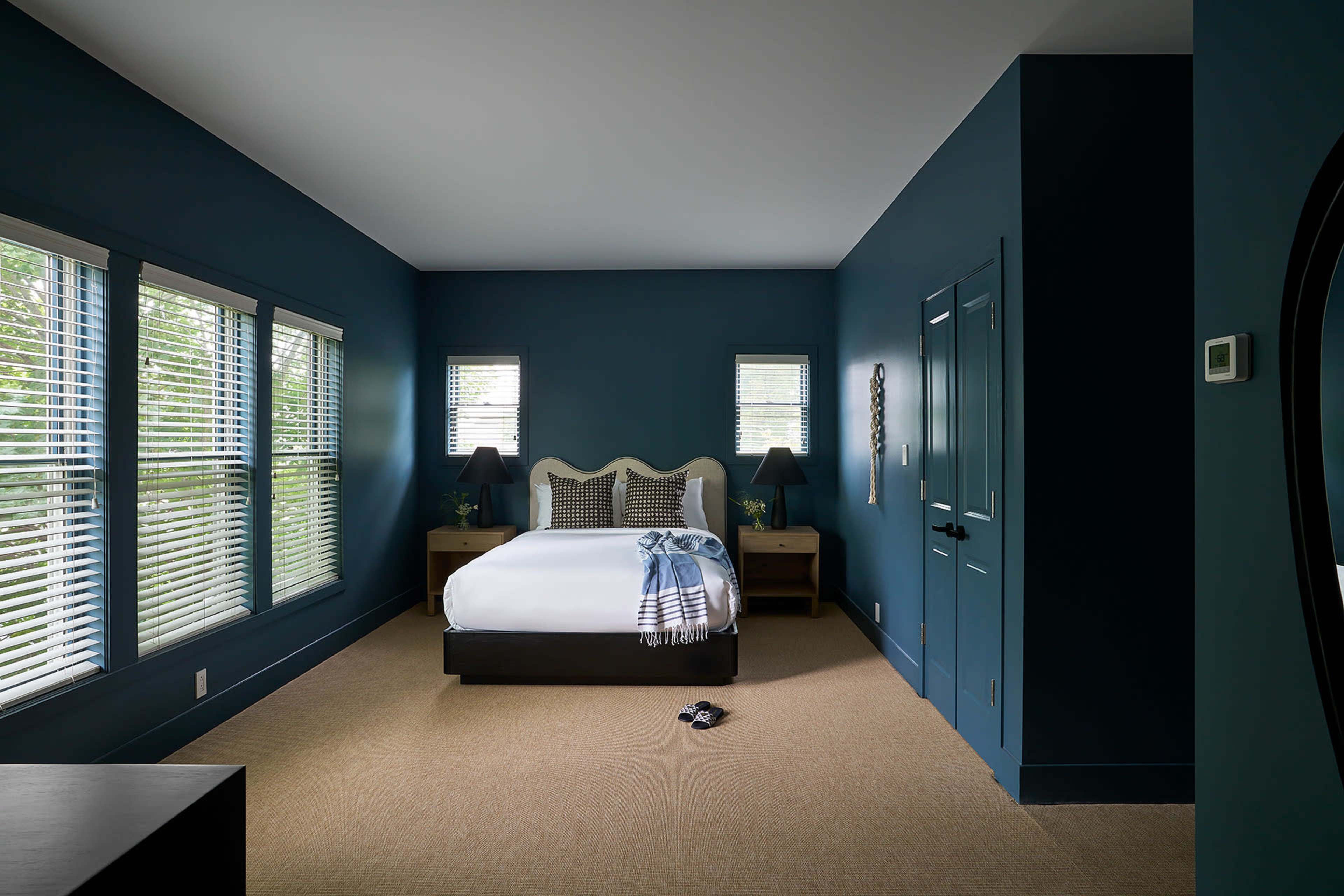 A bedroom with a blue wall, a queen-sized bed, two bedside tables with lamps, and window blinds.