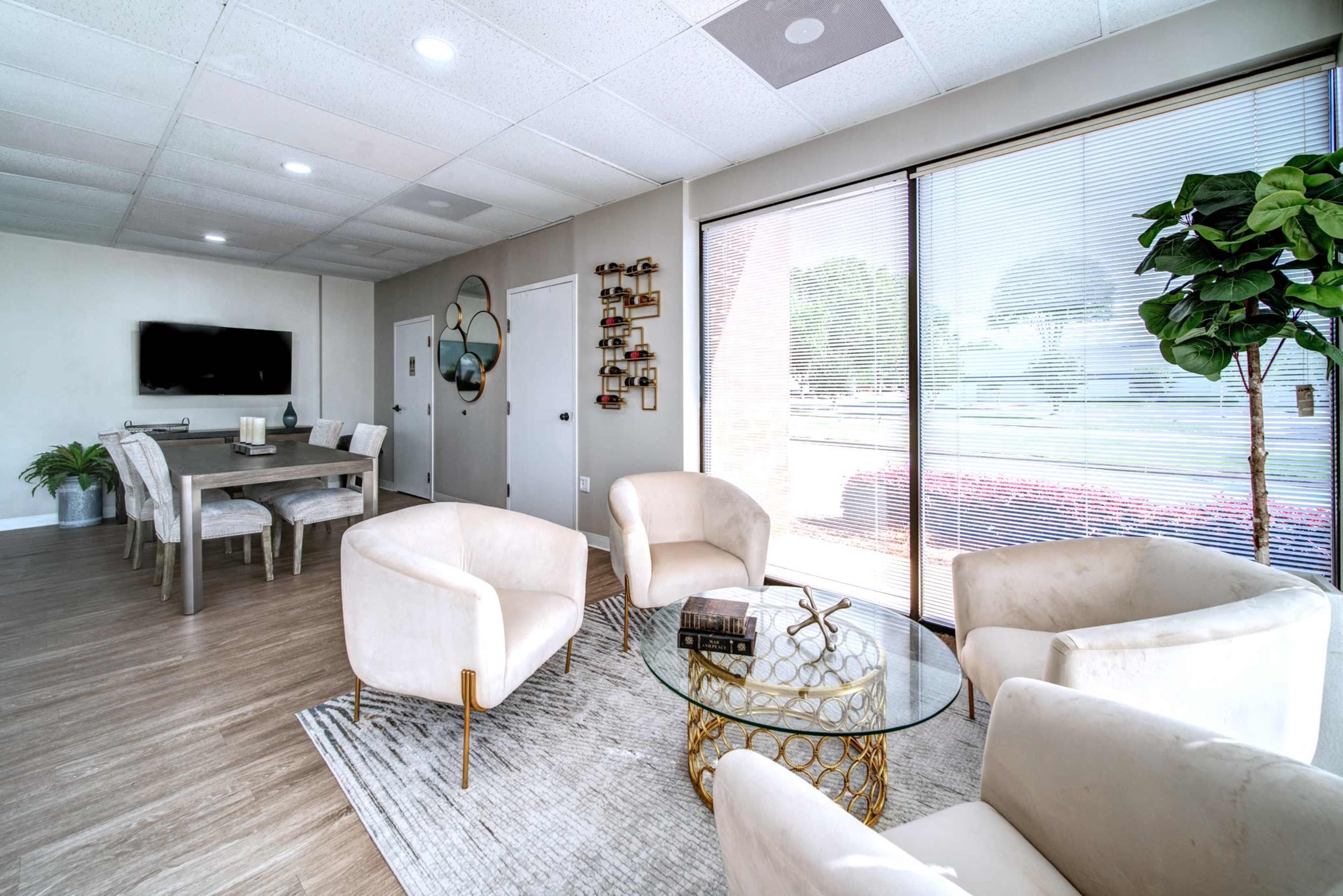 A modern living area featuring a round coffee table, four upholstered chairs, a dining table in the background, and large windows with blinds.
