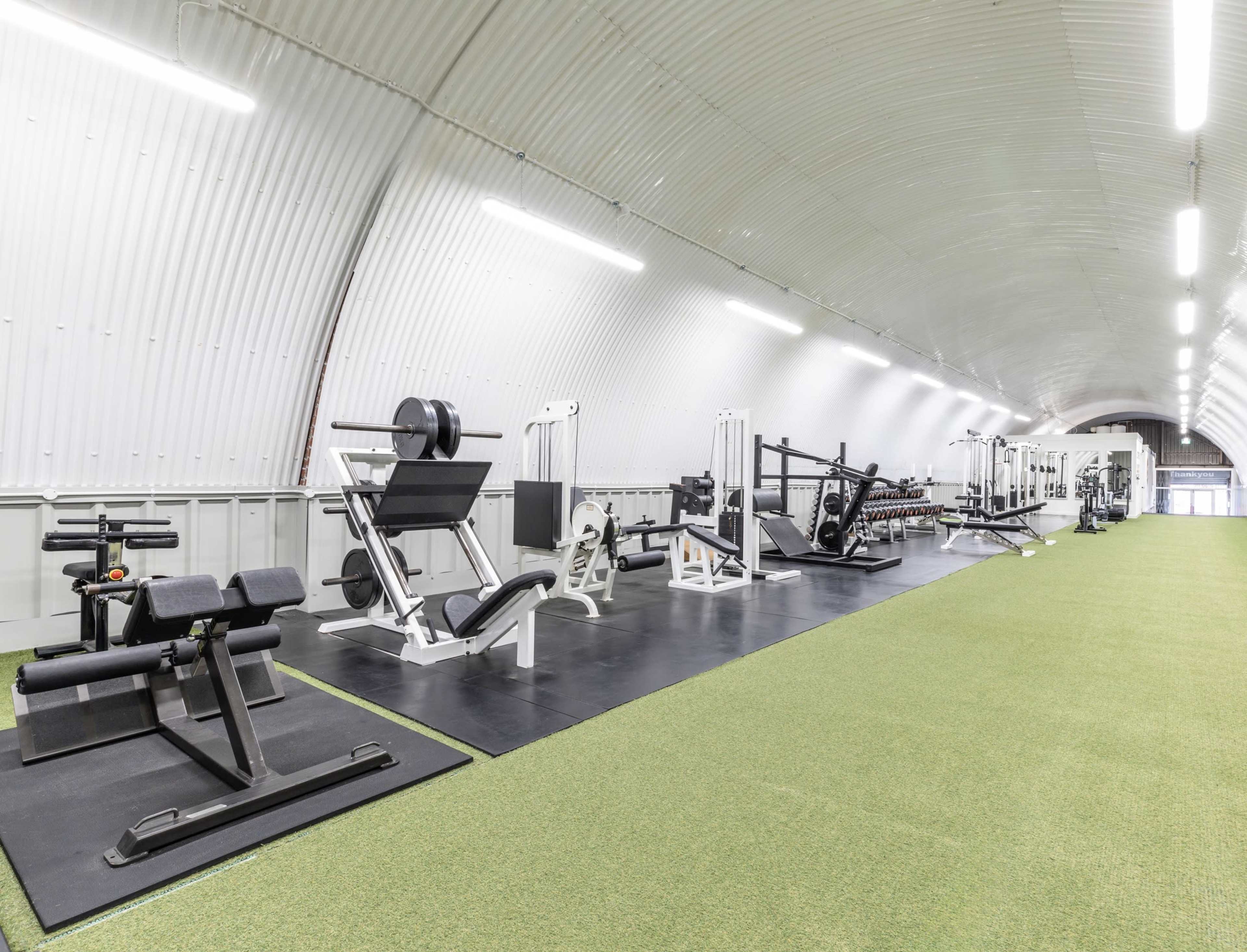 The image shows a long, narrow gym with various exercise equipment arranged along the walls and a green turf flooring in the center.