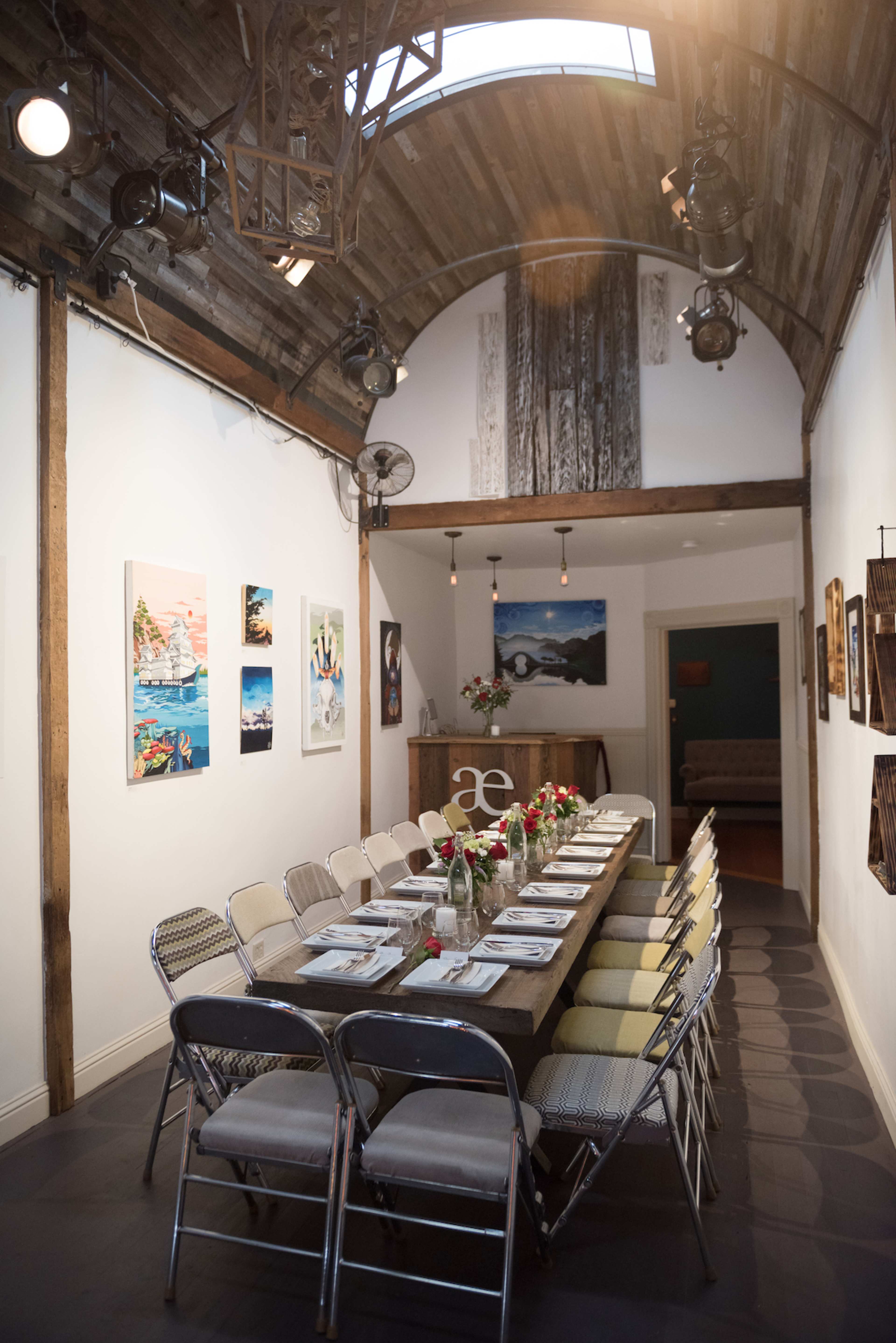A long dining table is set with flowers and chairs in a warmly lit, rustic room featuring artwork on the walls.