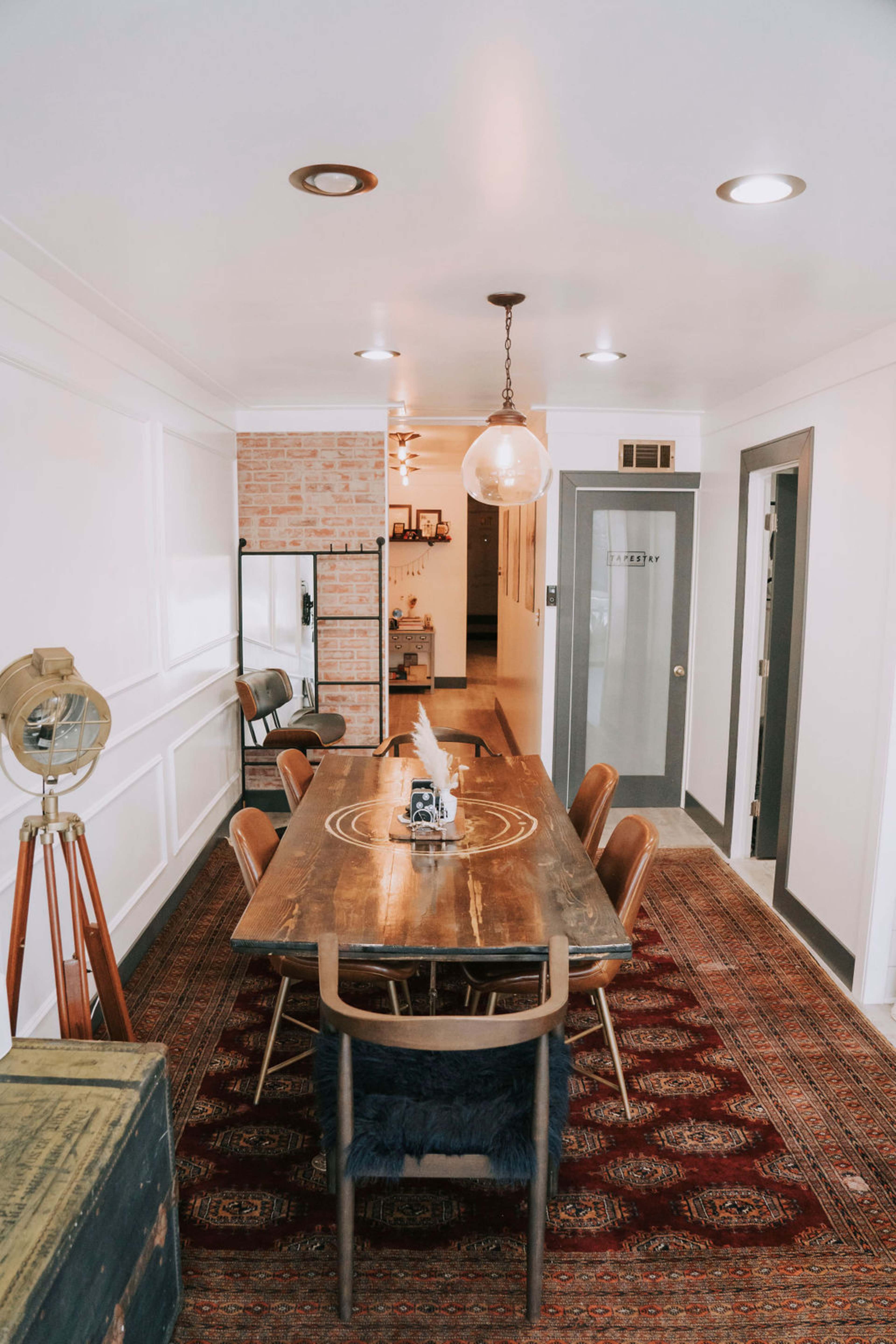 A dining area with a large wooden table surrounded by six chairs, set against a background of brick and white walls, with a rug on the floor and a hanging light fixture above.