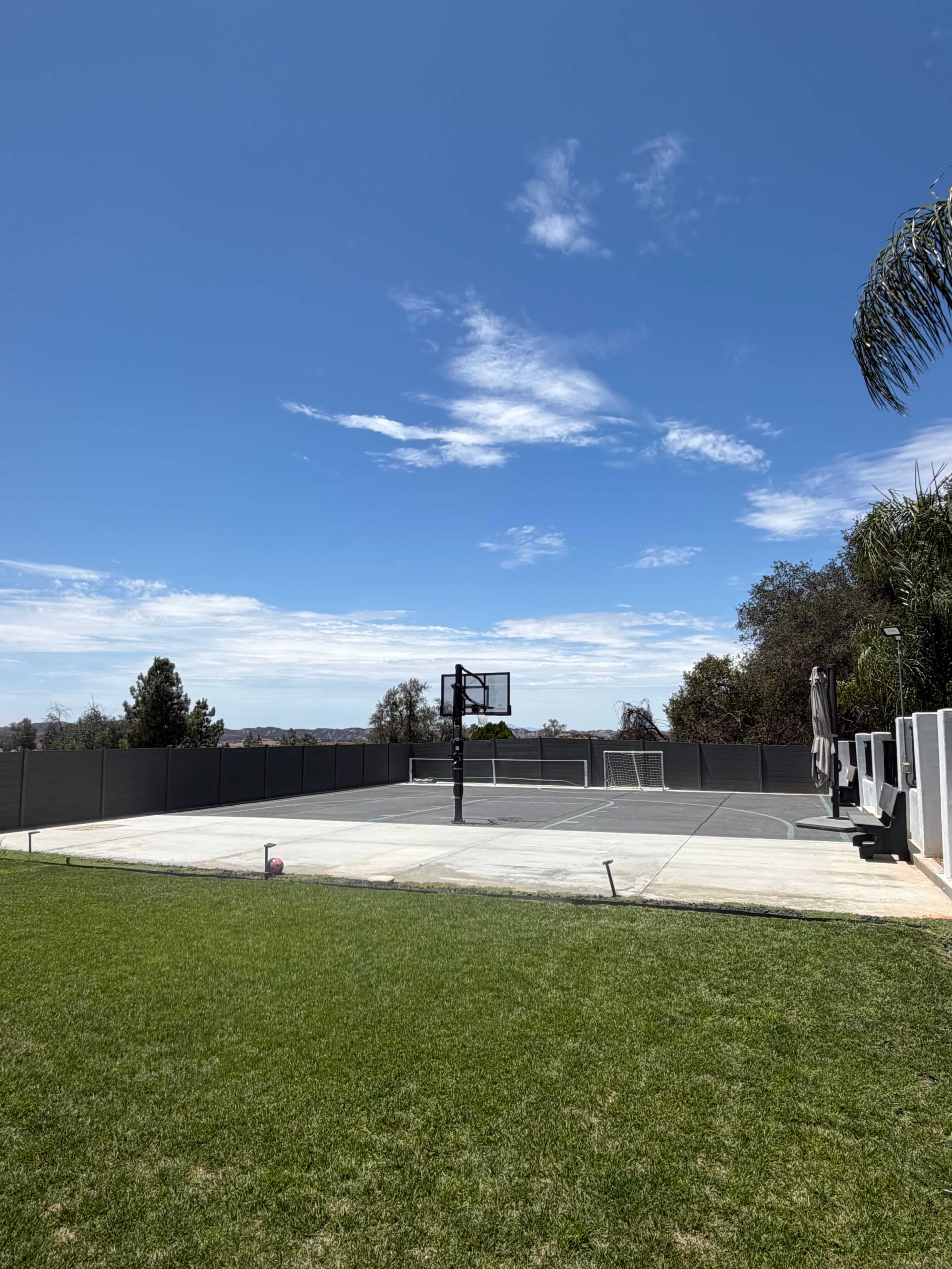 The image shows a basketball court with a hoop and a soccer goal in the background, surrounded by a grassy area under a clear blue sky.