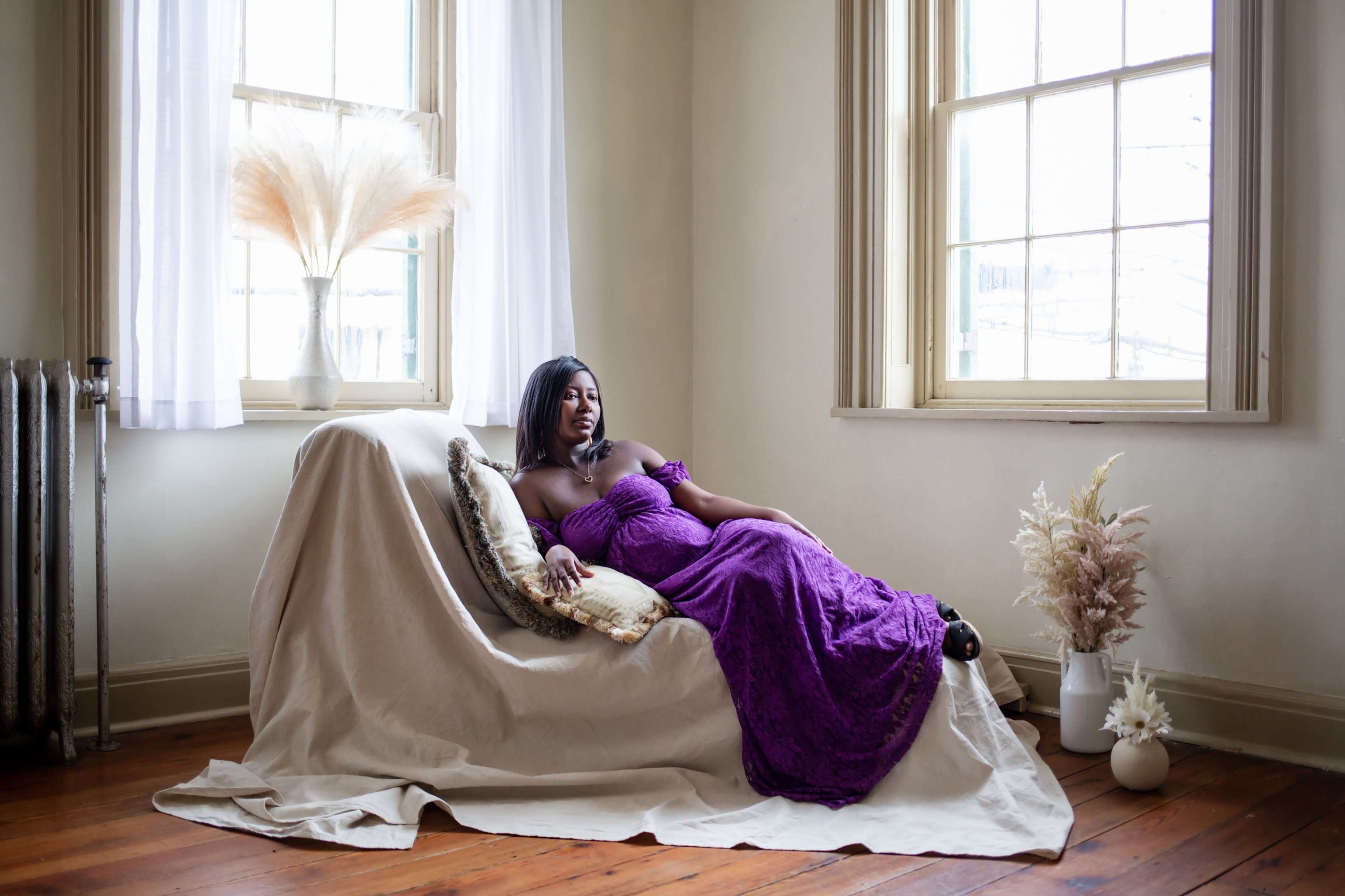 A person in a purple dress is reclining on a sofa in a room with large windows and decorative plants.
