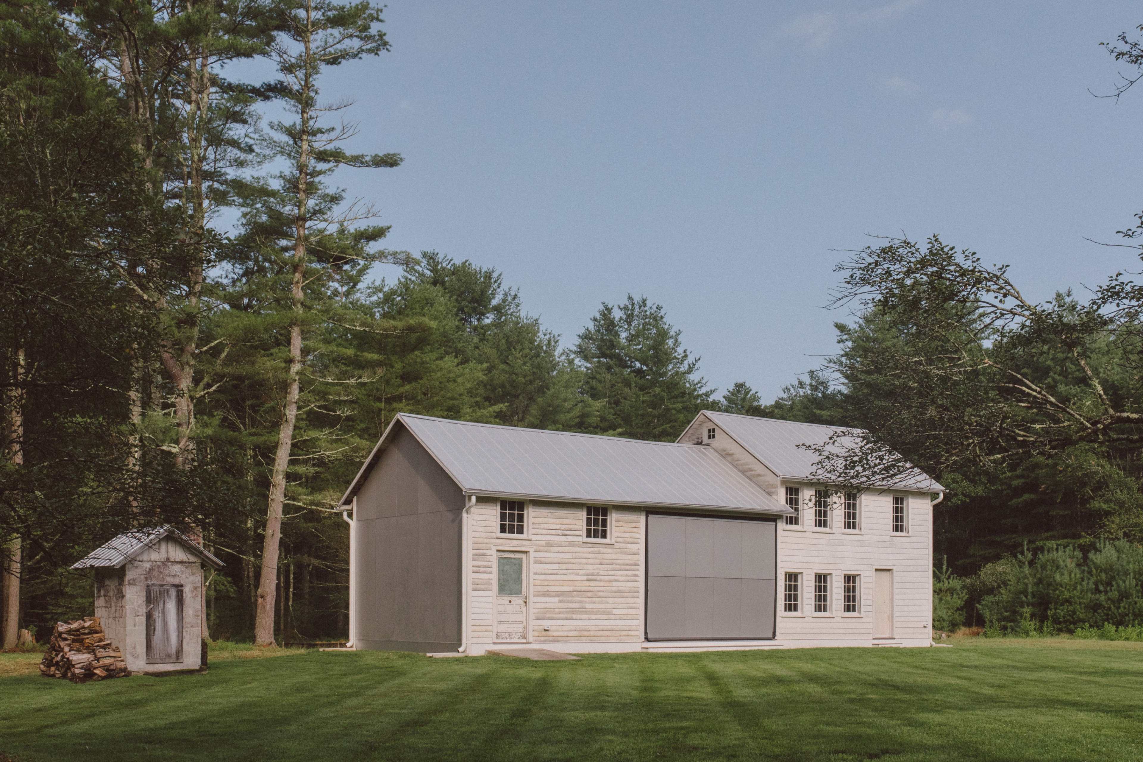 A rustic two-story house with a gray metal roof sits on a green lawn surrounded by tall trees, next to a small shed and a stack of firewood.