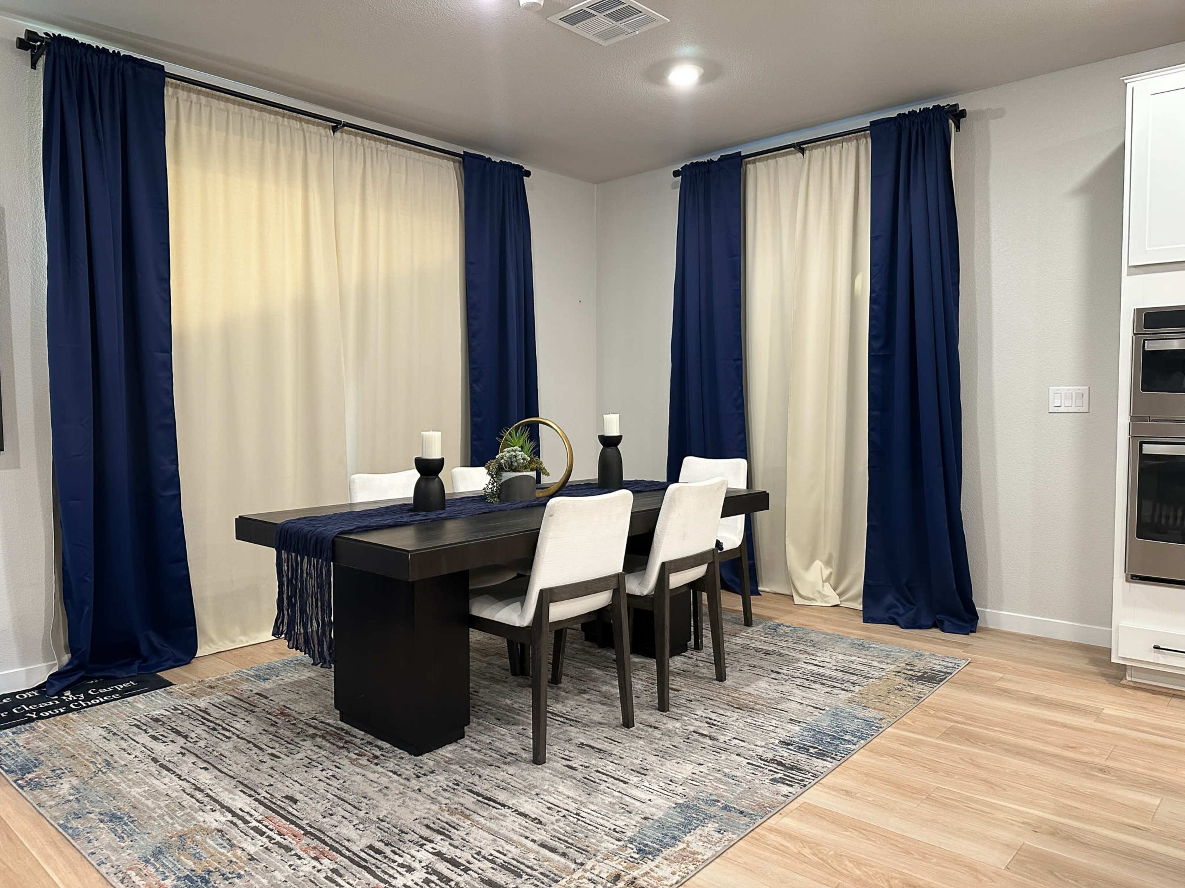 A dining area features a dark wooden table surrounded by four white chairs, with decorative candles on the table and navy blue curtains framing light-colored drapery on the windows.