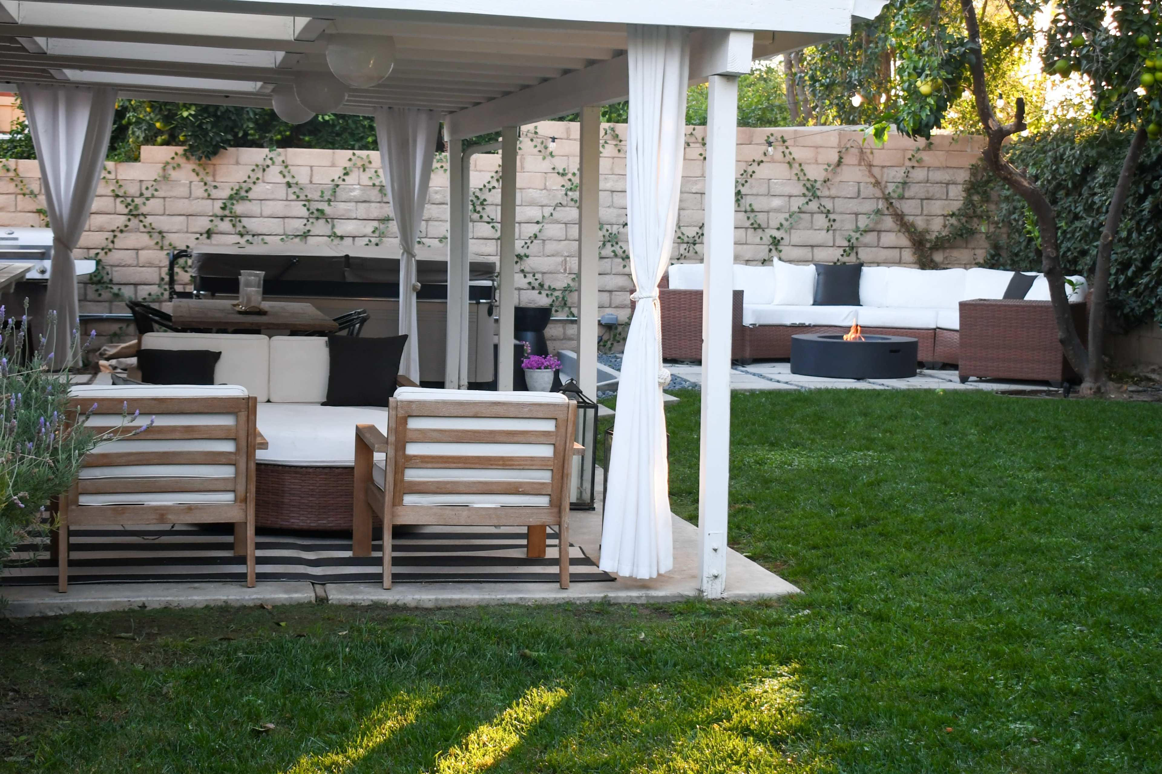 A well-designed backyard with a covered seating area featuring wooden chairs and a fire pit, surrounded by green grass and a sectional sofa.