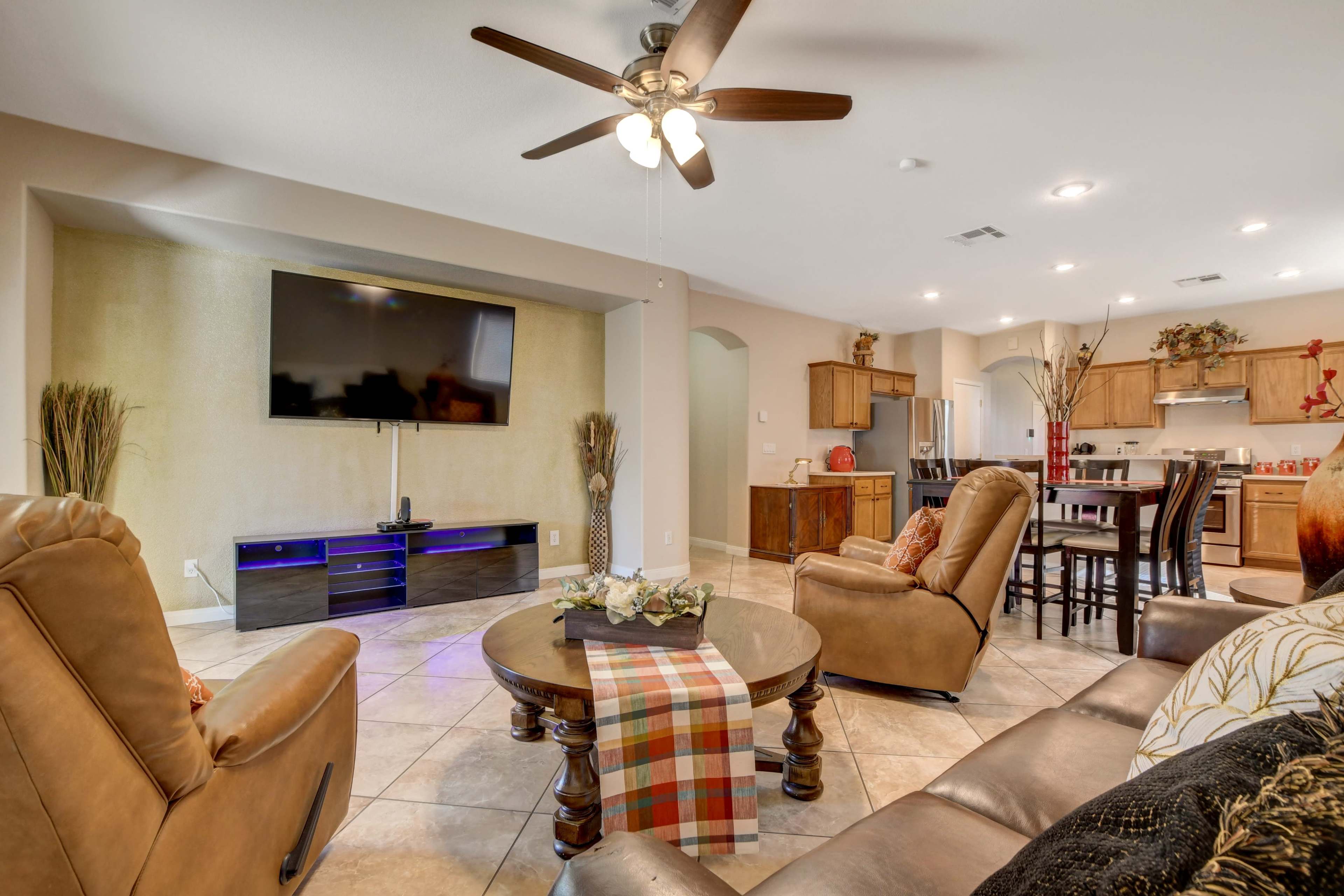 The scene shows a living room featuring a seating area with two armchairs and a coffee table, a wall-mounted television, and an open kitchen in the background.