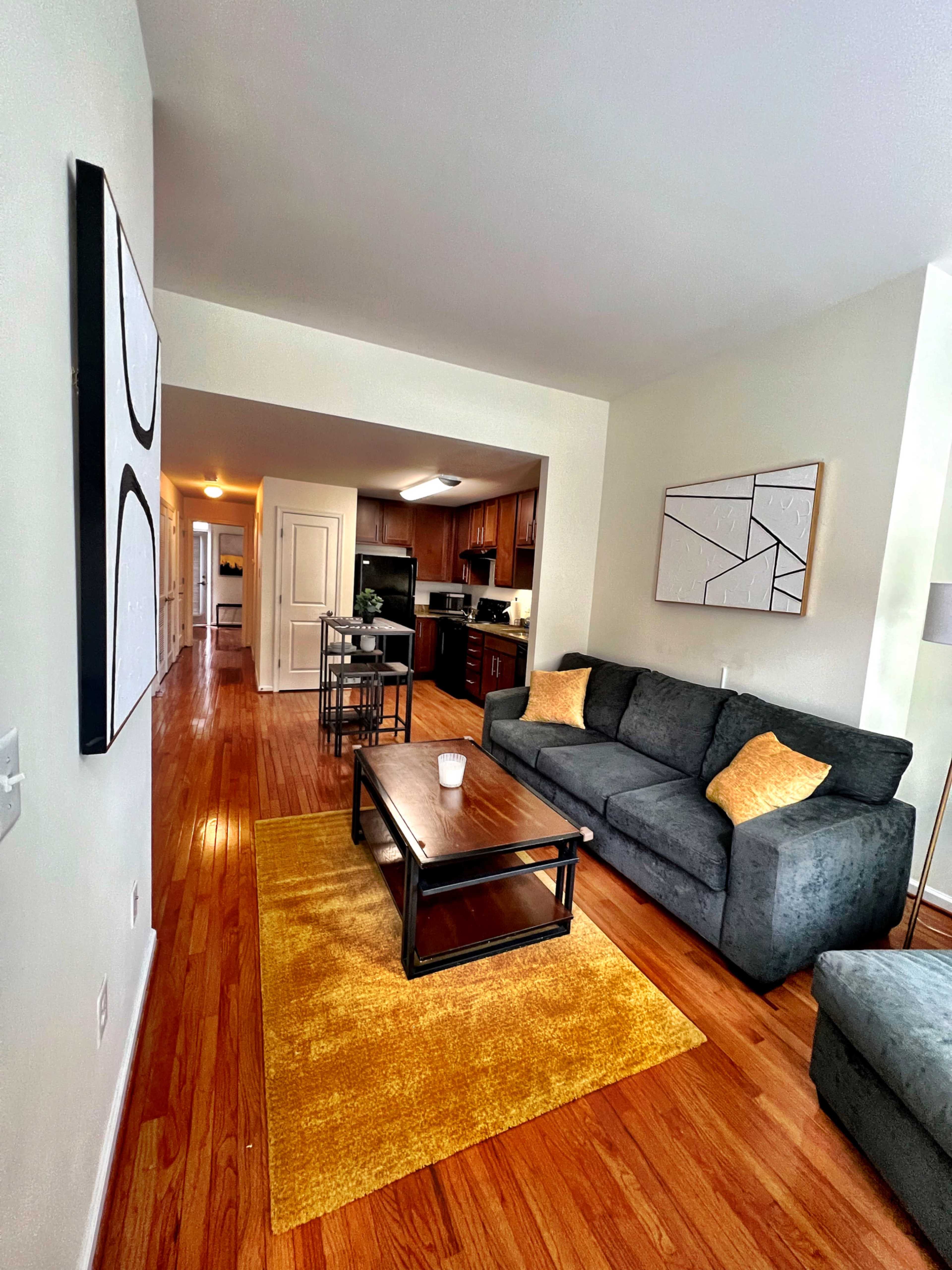 The image shows a living room with two gray sofas, a coffee table, a yellow rug, and an open kitchen in the background.
