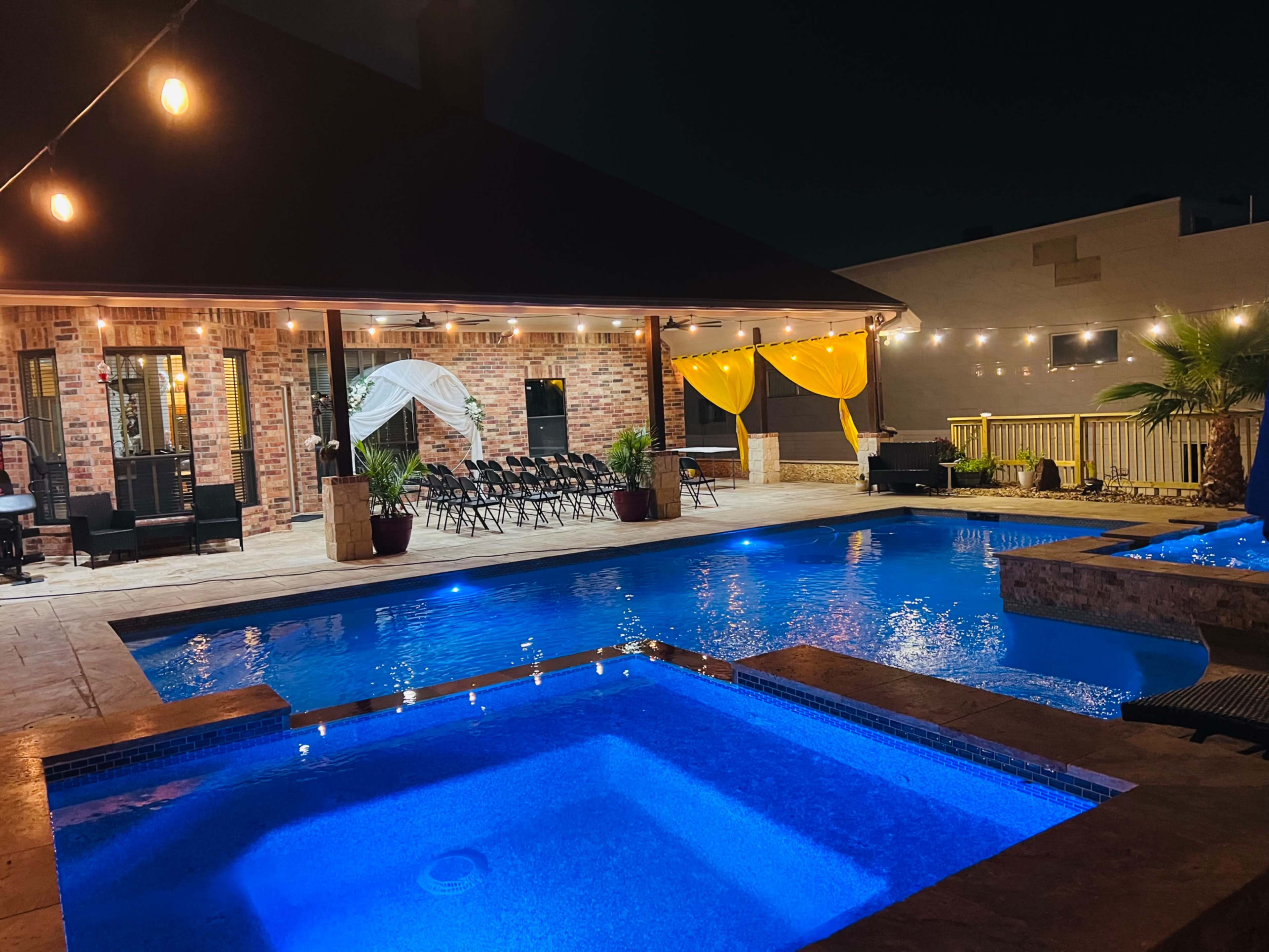 The image shows a well-lit outdoor pool area with a hot tub, surrounded by lounge chairs and decorated with string lights and yellow drapes.