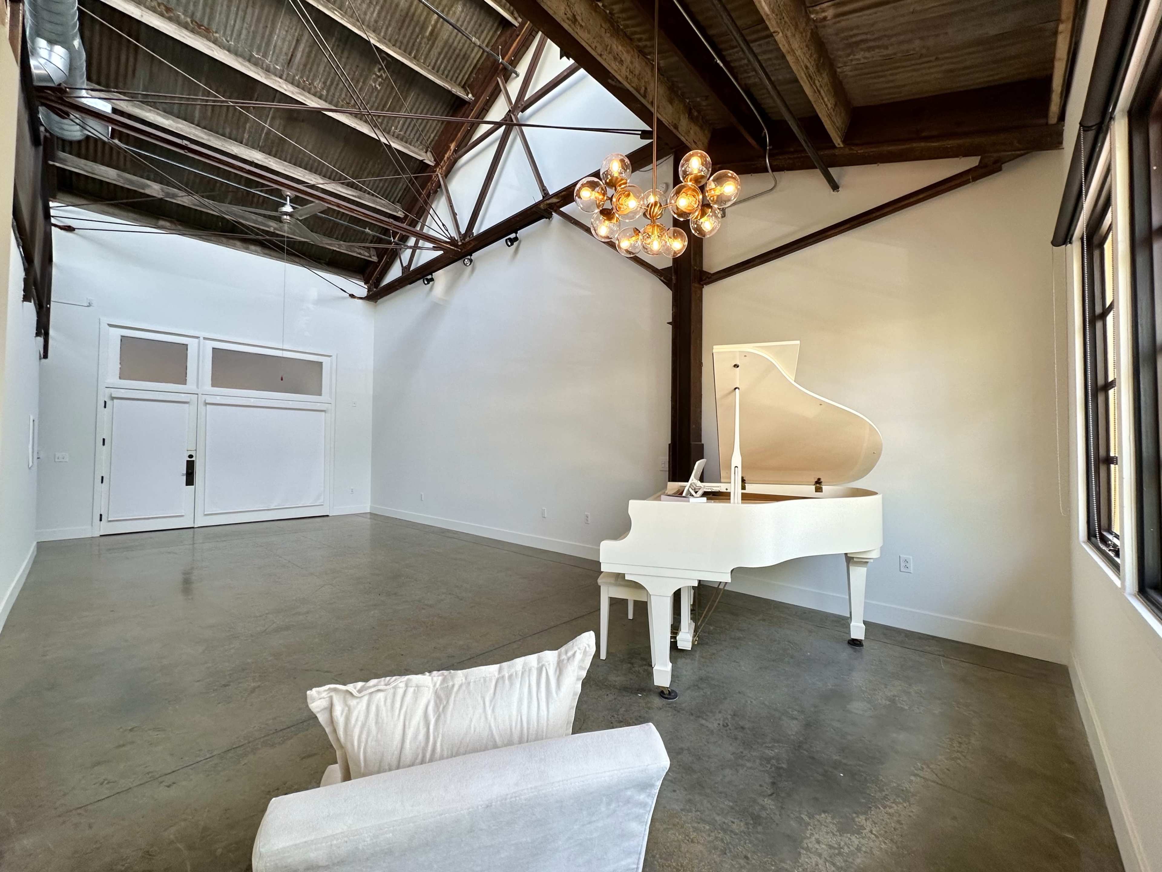 A spacious room with a white grand piano, a light fixture with multiple bulbs, and a single white sofa in the corner, featuring exposed beams and concrete flooring.