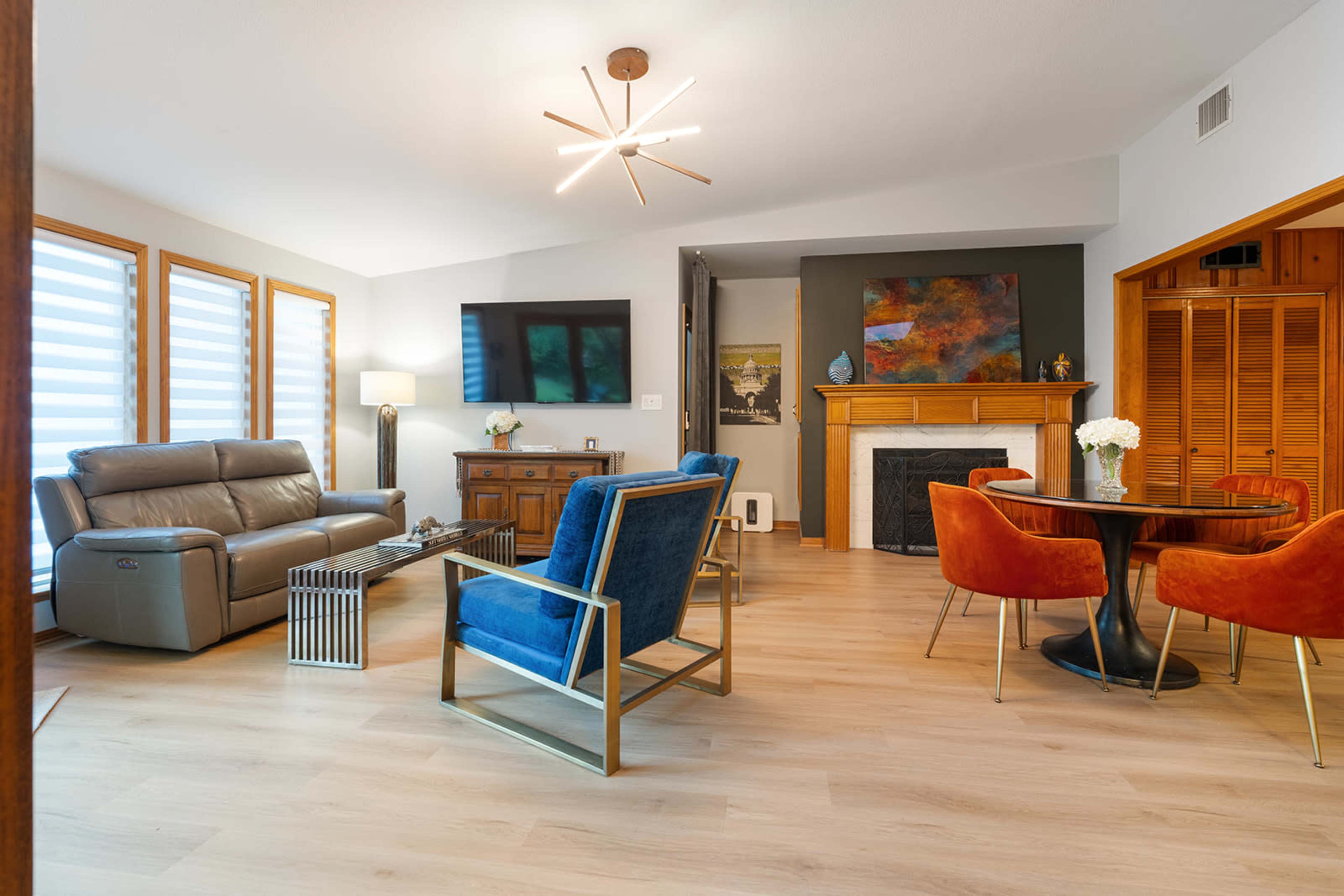 The image shows a modern living room with a combination of a gray sofa, blue chairs, a wooden television stand, and a round dining table set against light-colored flooring and large windows.