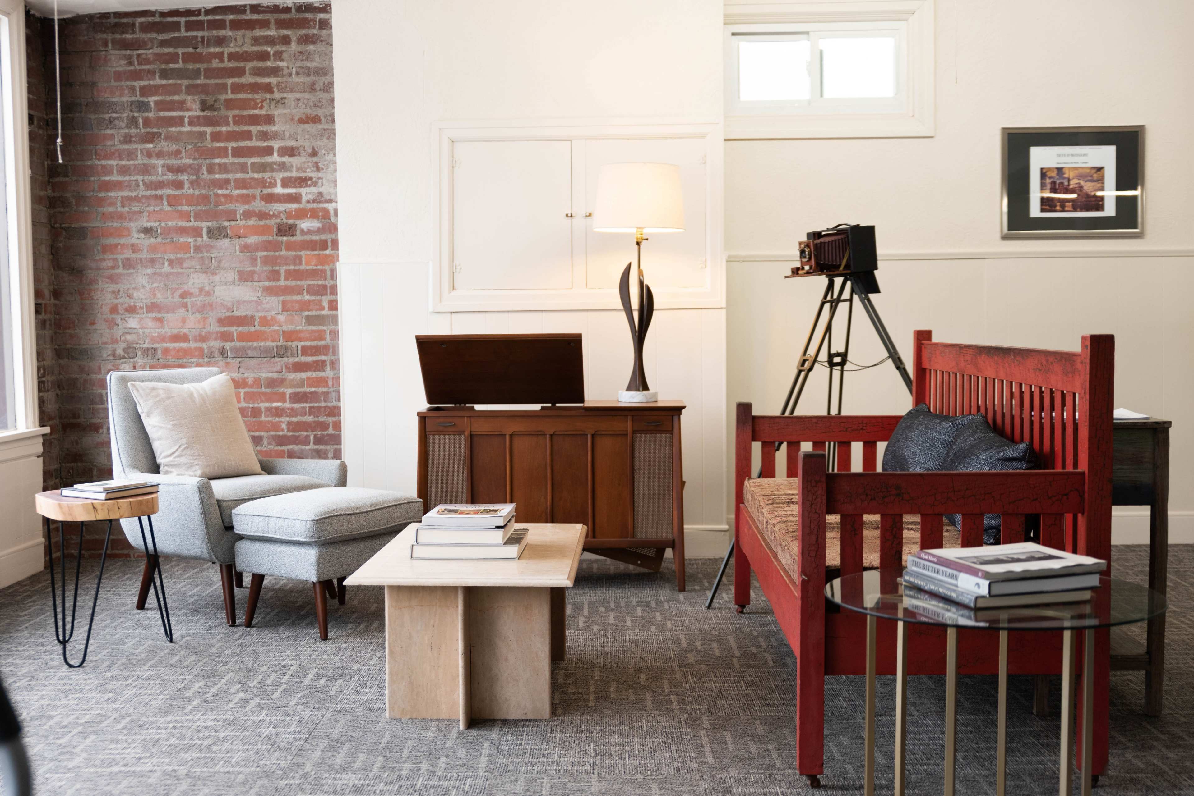 A modern living space featuring a mix of vintage and contemporary furniture, including a red wooden bench, a gray armchair, a wooden cabinet, and a tripod camera.