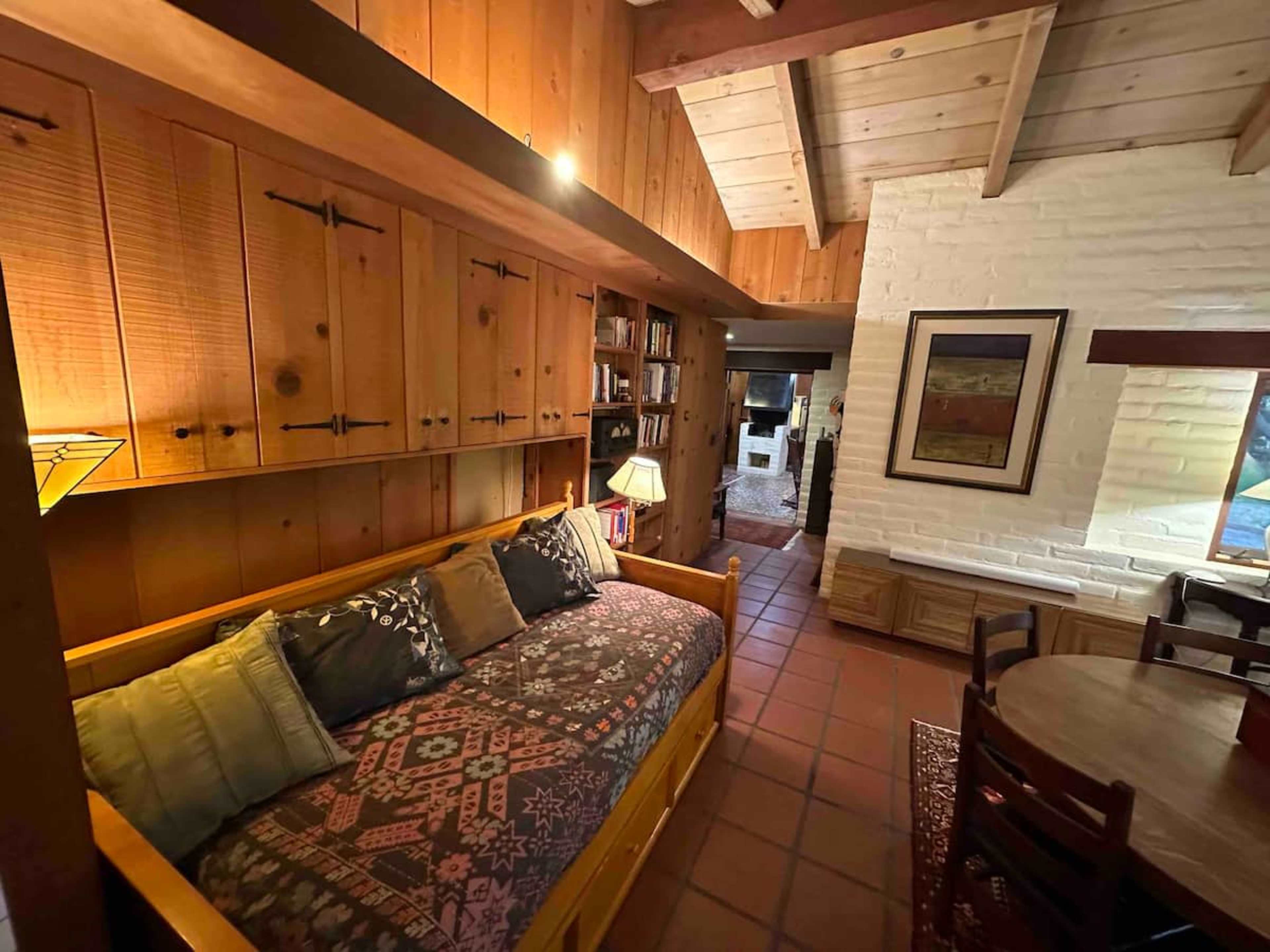 A cozy living space featuring a wooden daybed, built-in cabinets, and a dining area, with exposed wooden beams and a brick wall in the background.
