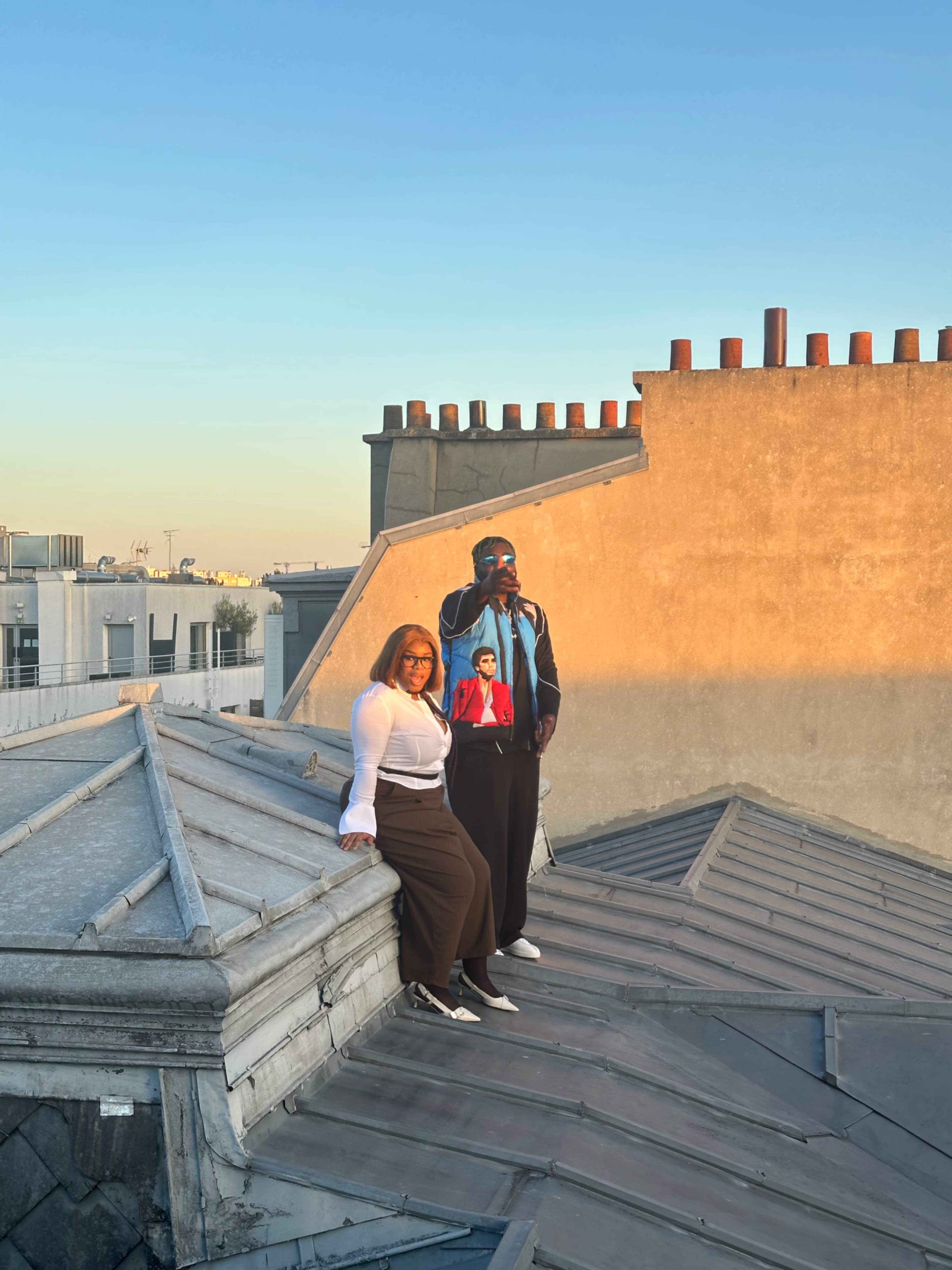 Two people are standing on a rooftop with a clear sky in the background, one wearing a white top and brown pants, and the other in a colorful jacket, as they pose for a photo.