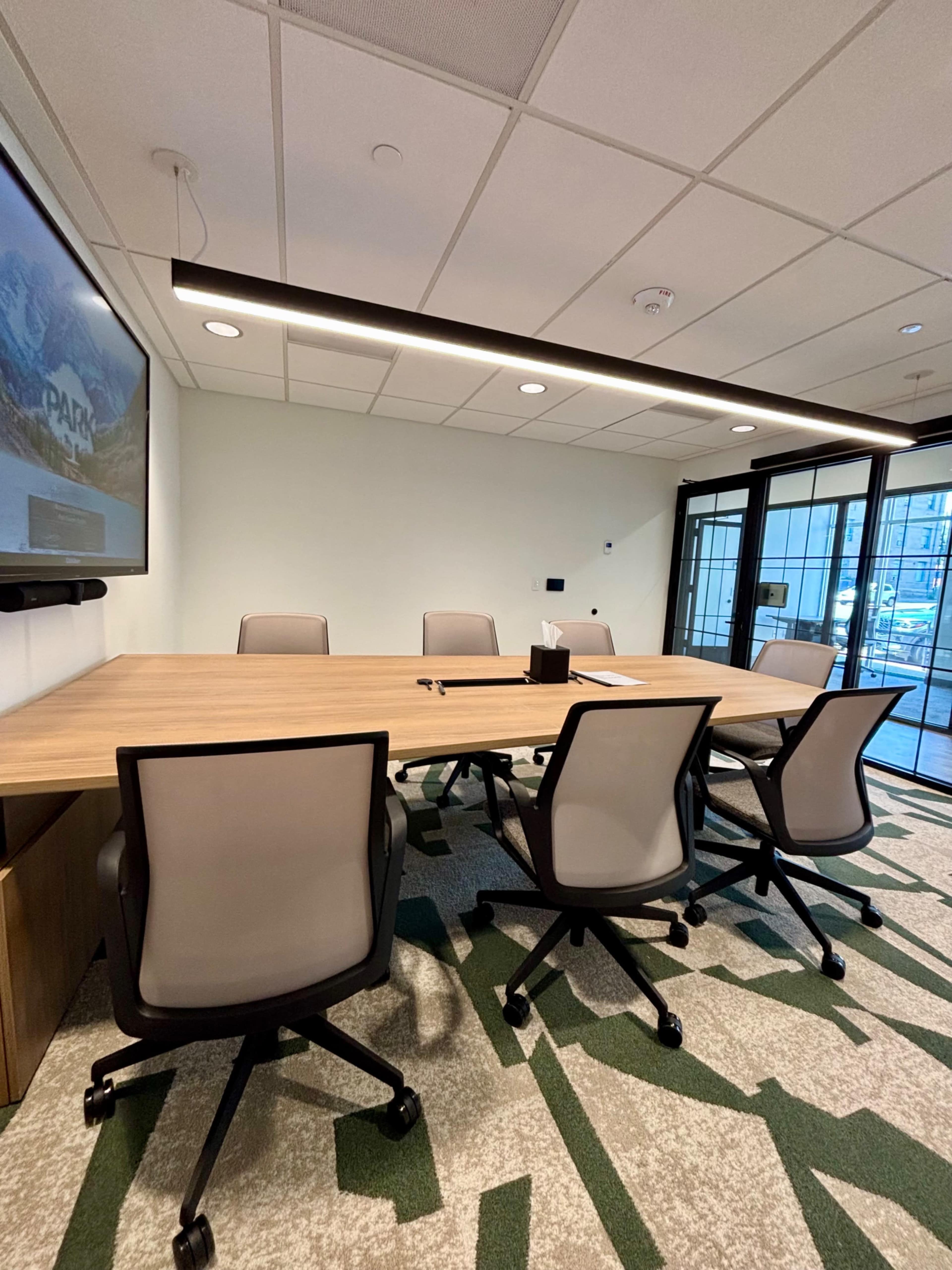 A conference room is set up with a long wooden table and five rolling chairs, featuring a large screen on one wall and large windows providing natural light.
