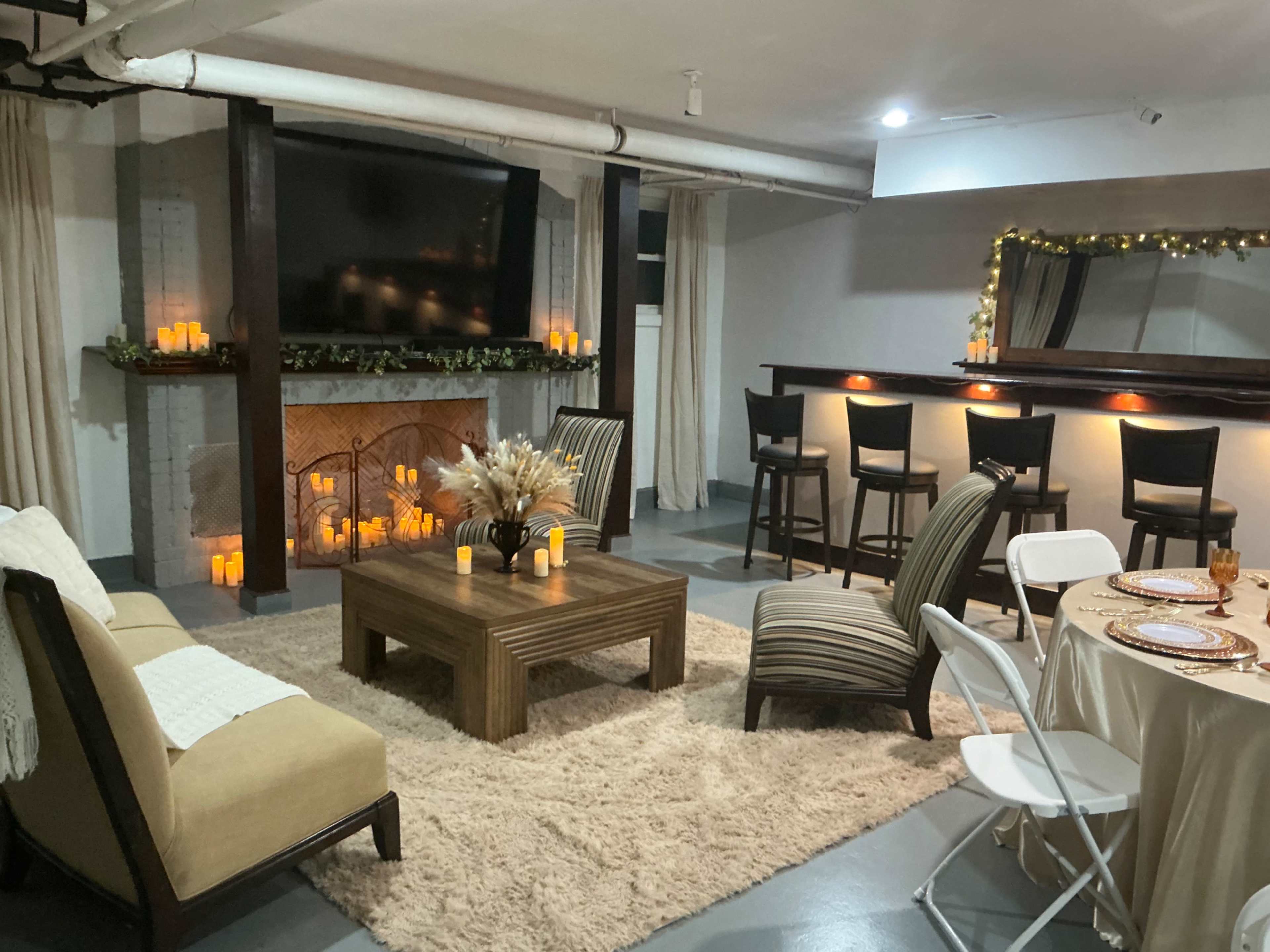 The image shows a cozy lounge area with a fireplace, a large TV, comfortable seating, a coffee table, and a bar with stools in a softly lit room.