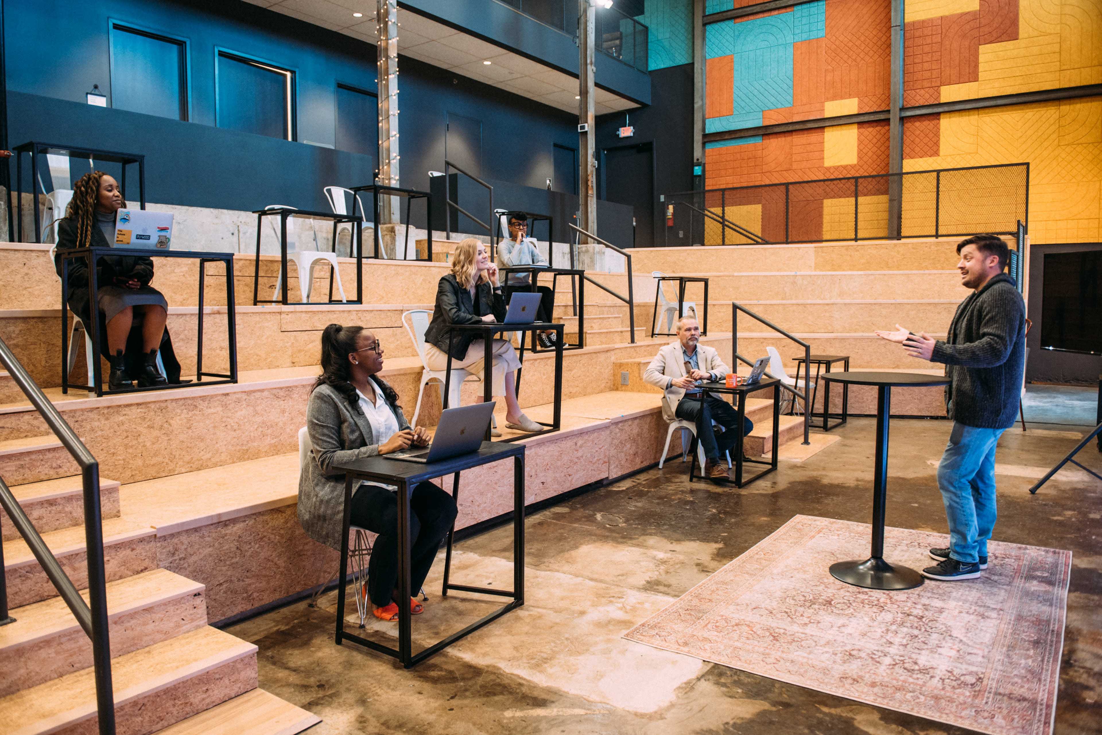 A man stands at a podium addressing a group seated at tables on a tiered wooden stage in a brightly colored, modern interior space.