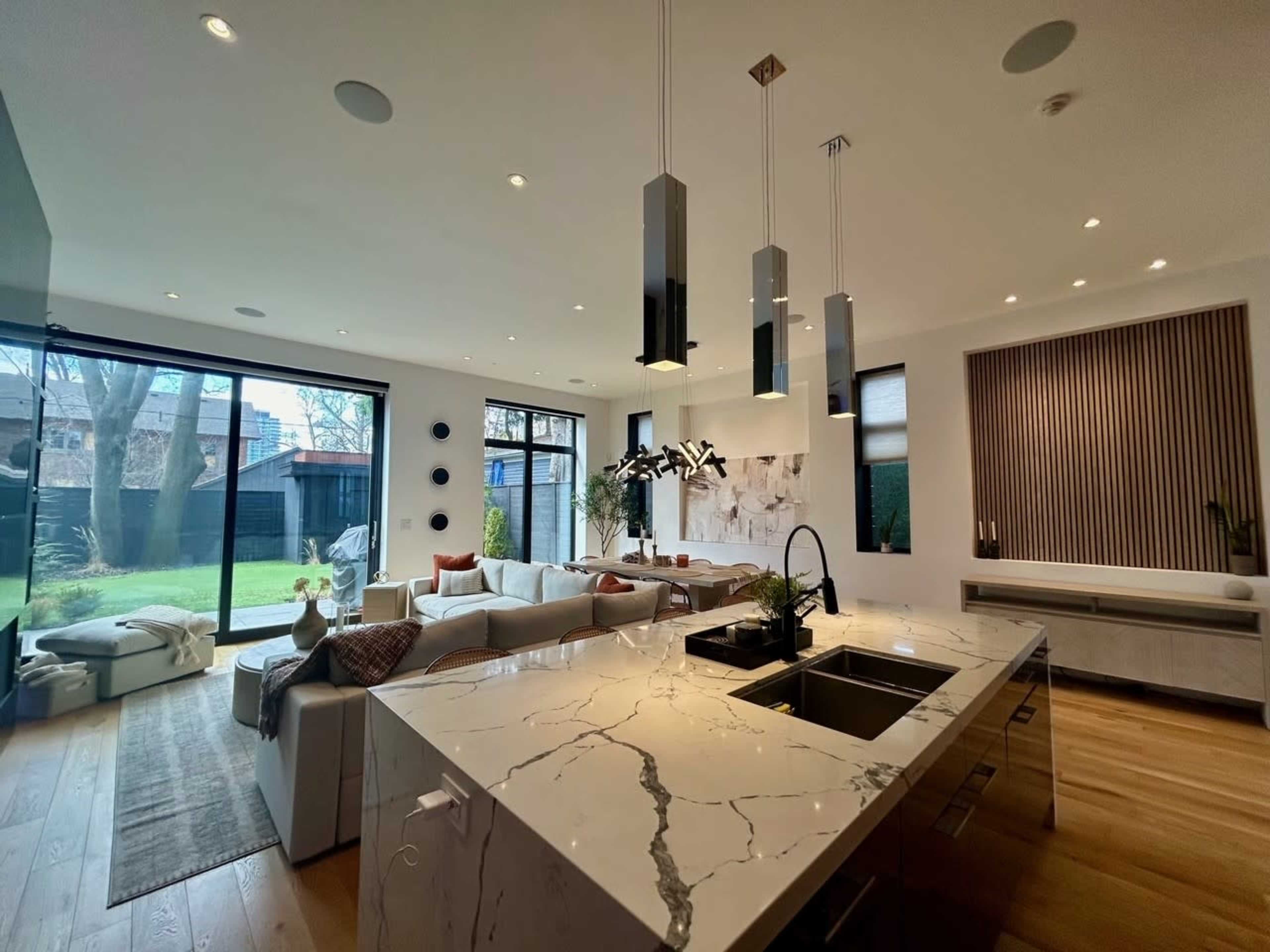 A modern living room features large windows overlooking a green space, with an open kitchen island made of marble and contemporary lighting fixtures.