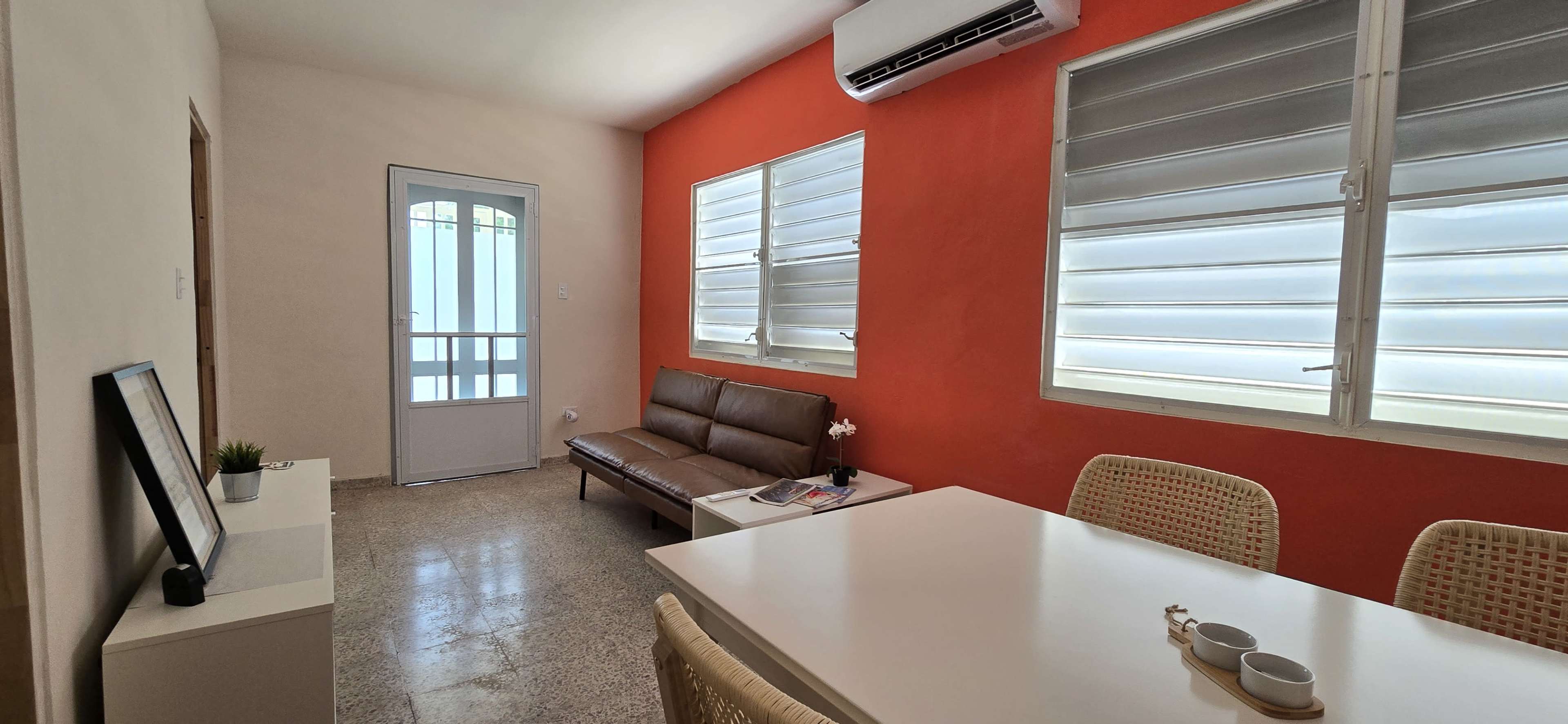 The image shows a minimalist room with a brown sofa, a white table, and large windows, featuring one wall painted in orange.