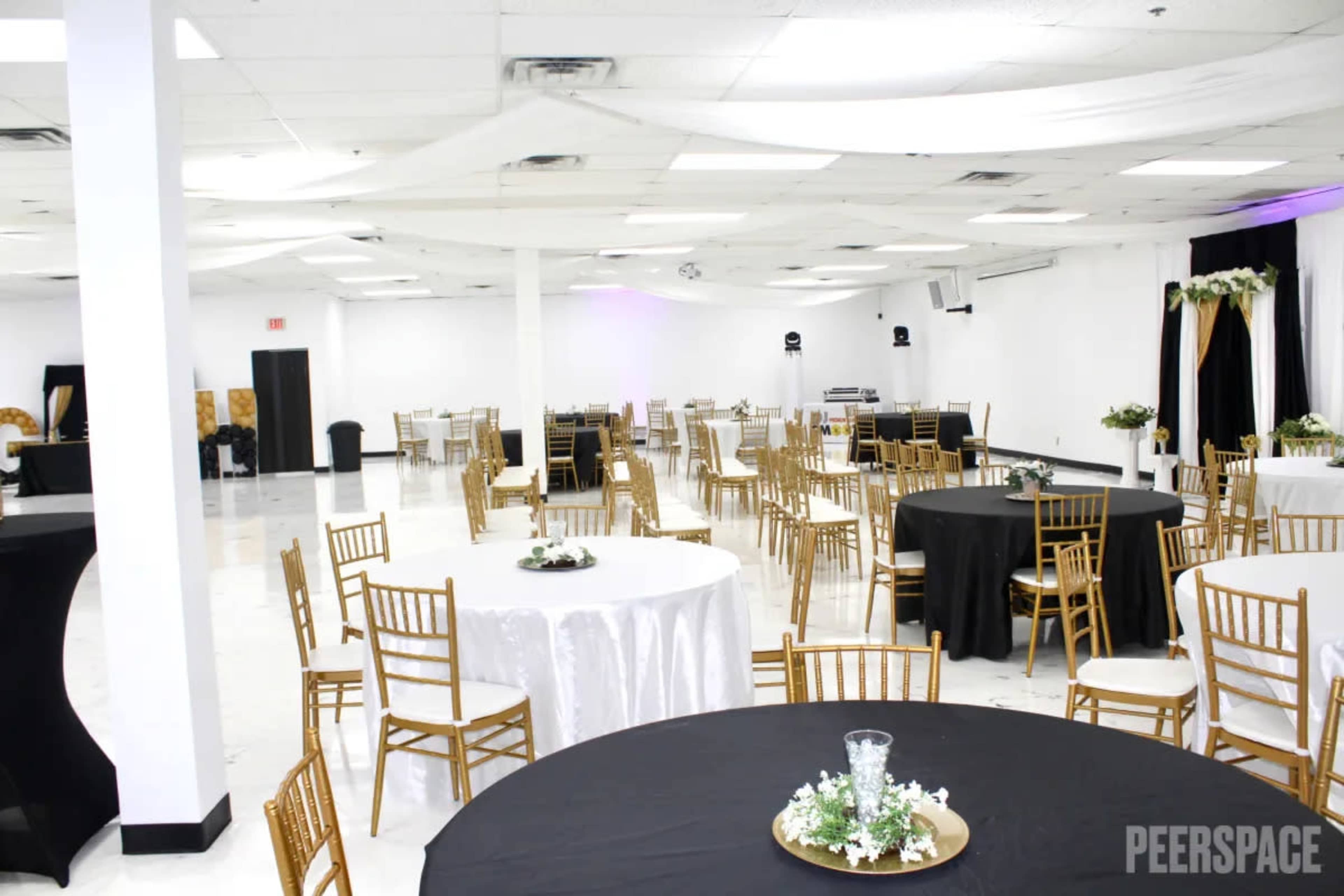 A spacious event hall features multiple round tables with gold chairs, arranged around a central area designed for gatherings.