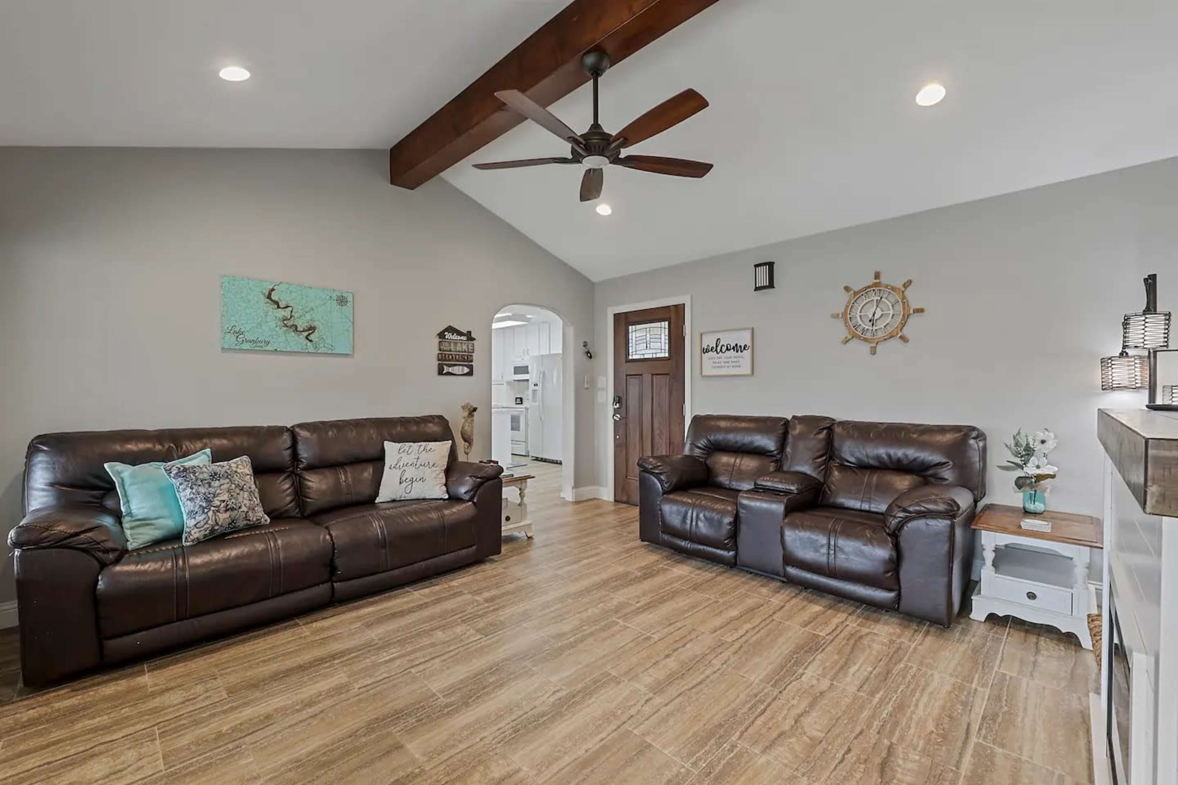 The living room features two brown leather couches, a wooden ceiling beam, and a light-colored wall decorated with art and a clock.