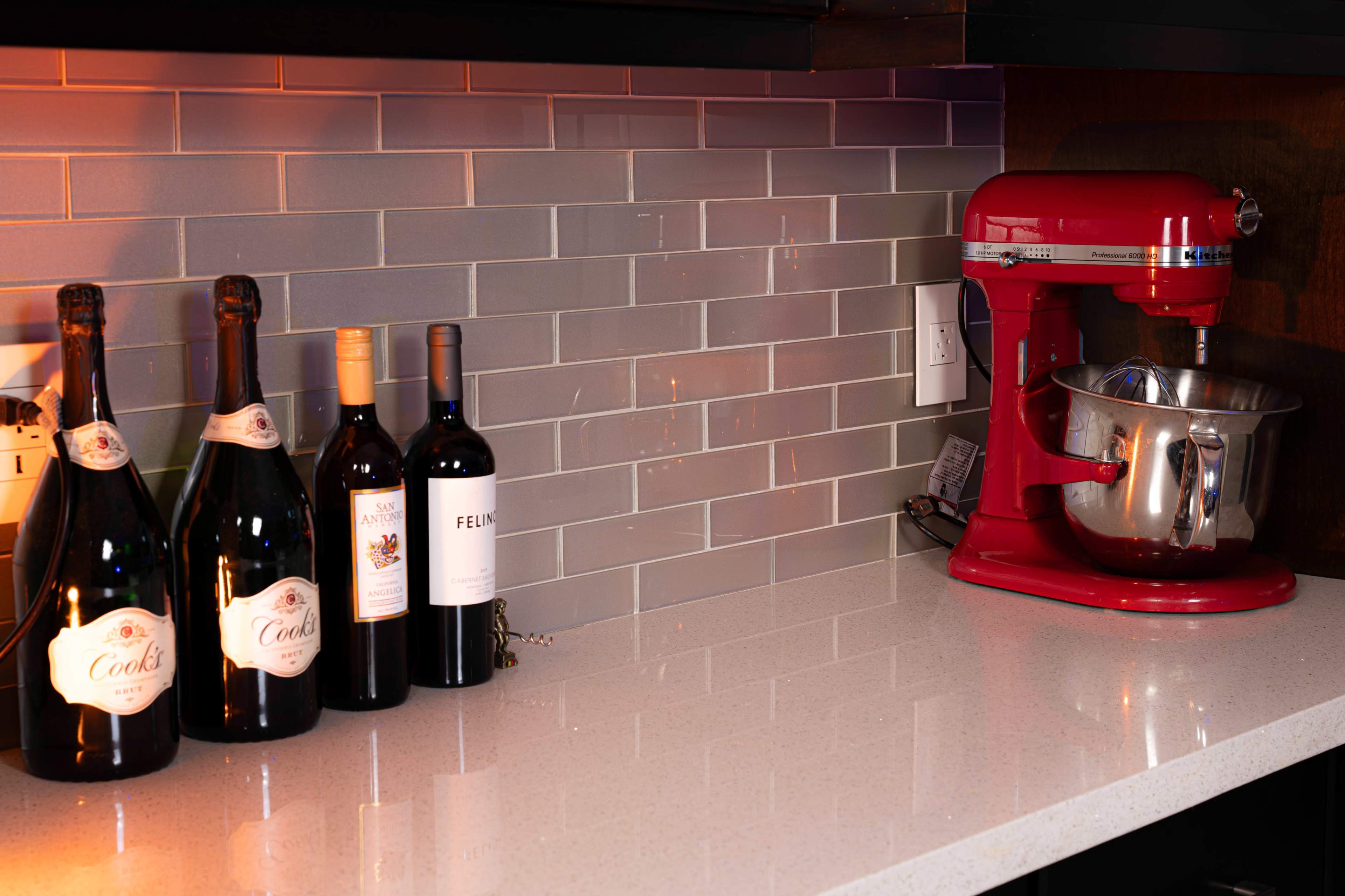 The kitchen countertop features several bottles of wine alongside a red stand mixer on a gray tiled backdrop.