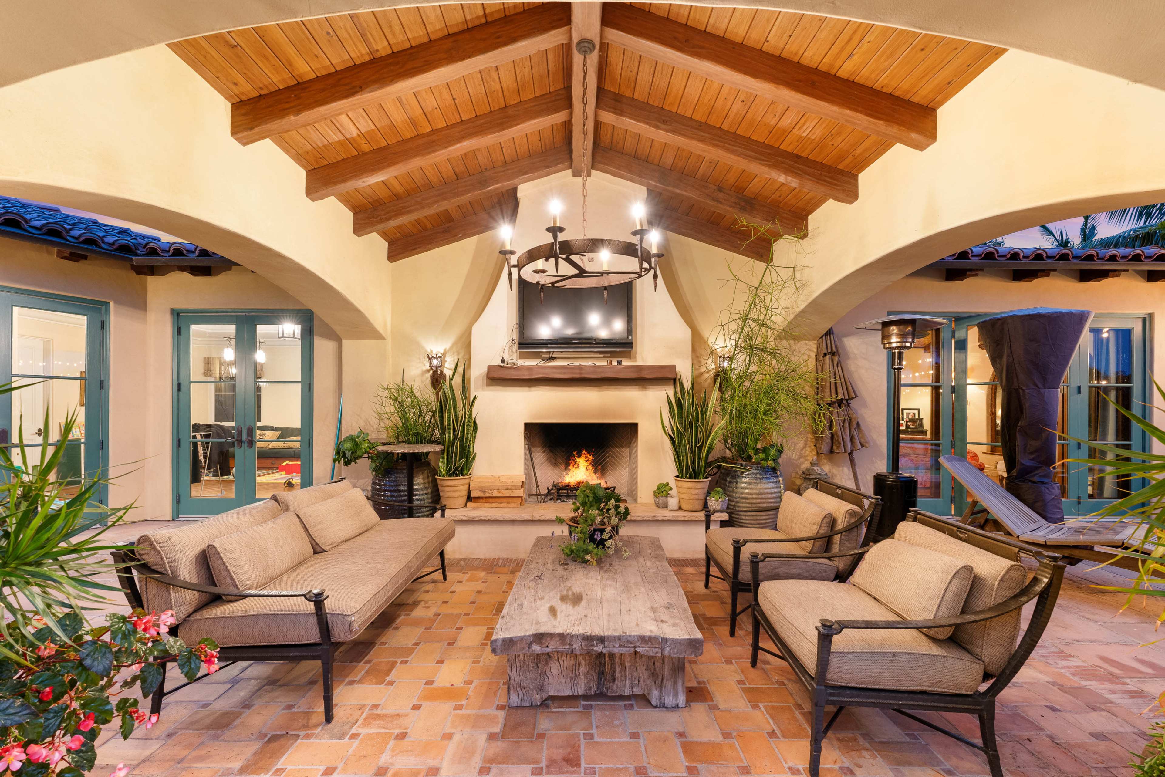 A cozy outdoor seating area with a wooden ceiling, a central fireplace, a large television mounted above the mantel, and potted plants enhancing the space.
