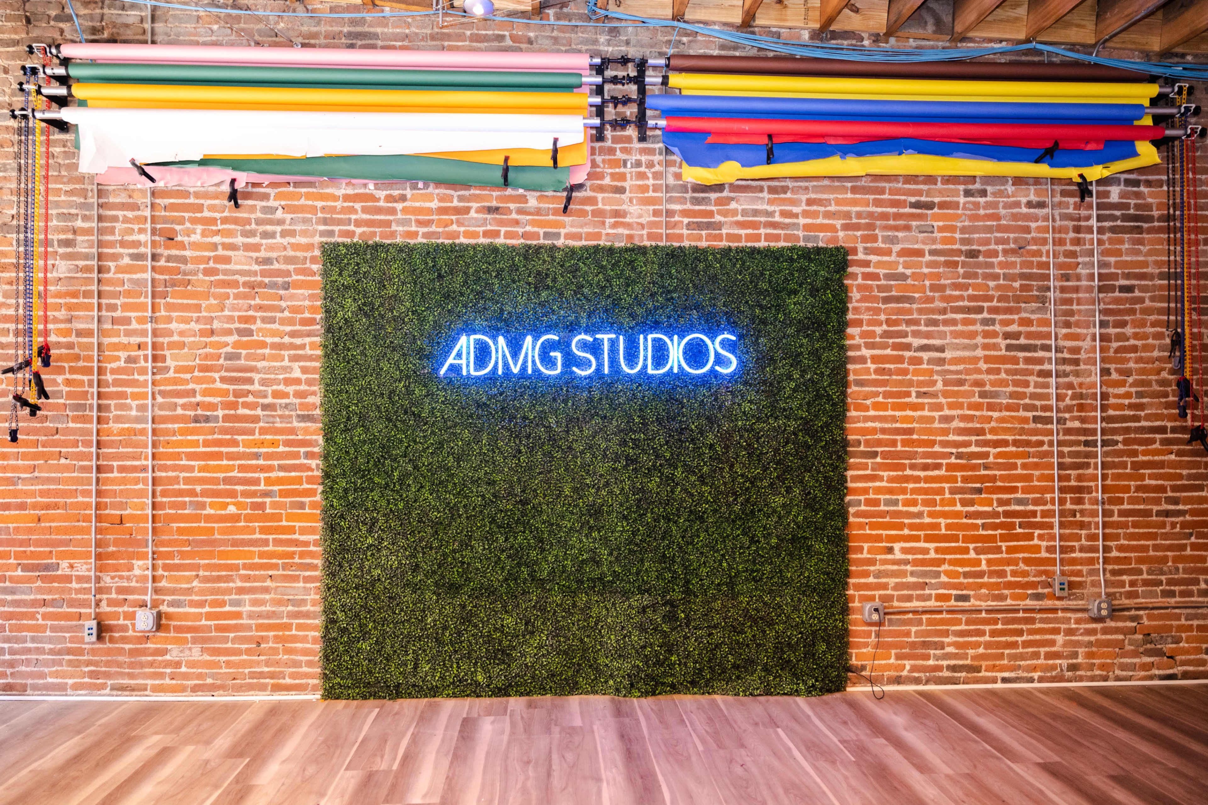 The image shows a brick wall with a green moss-like backdrop and the neon sign "ADMG STUDIOS" illuminated above it, complemented by colorful rolls of backdrops hanging overhead.