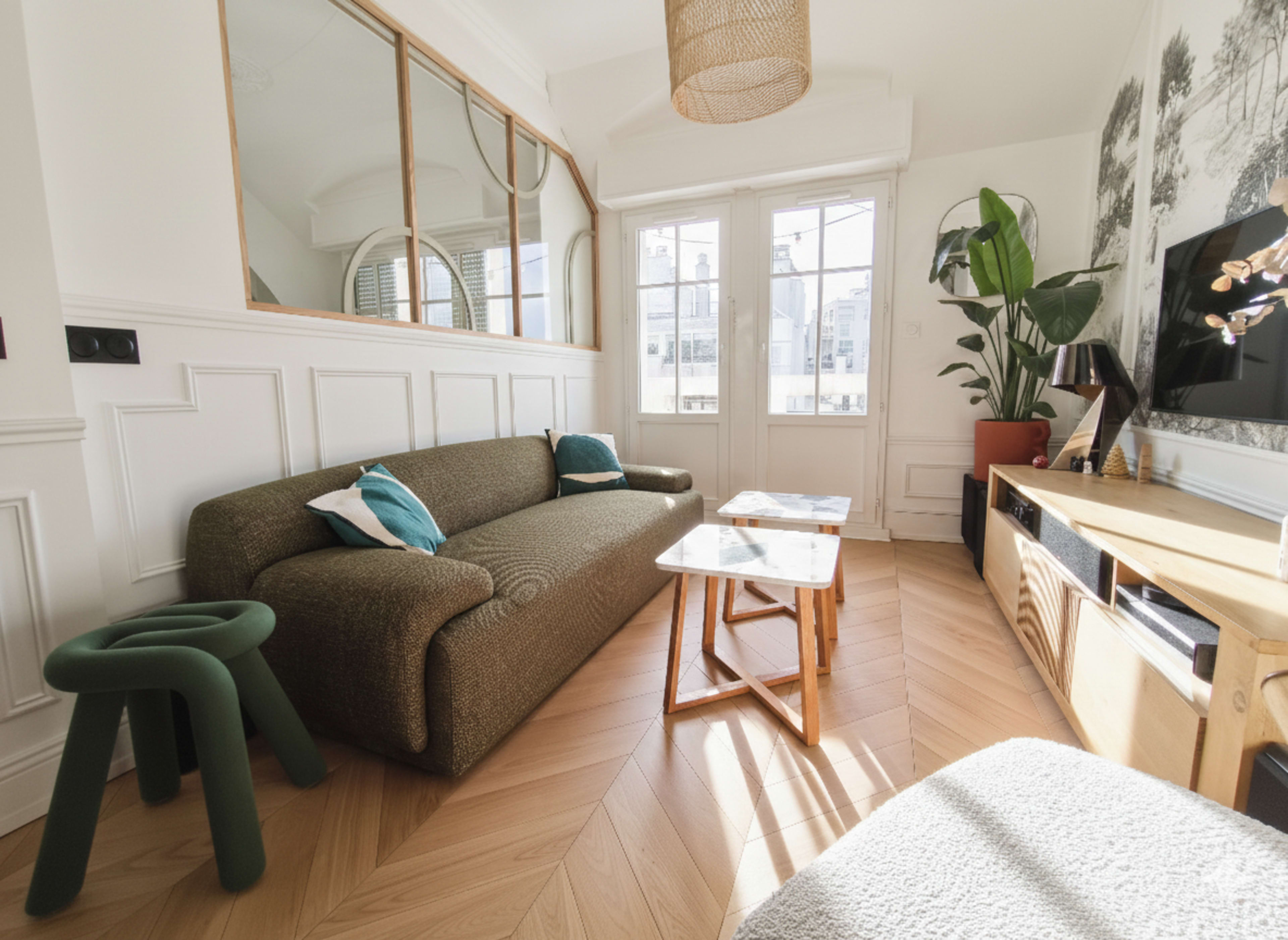 The living room features a textured sofa, wooden side tables, and large windows that allow natural light to fill the space.