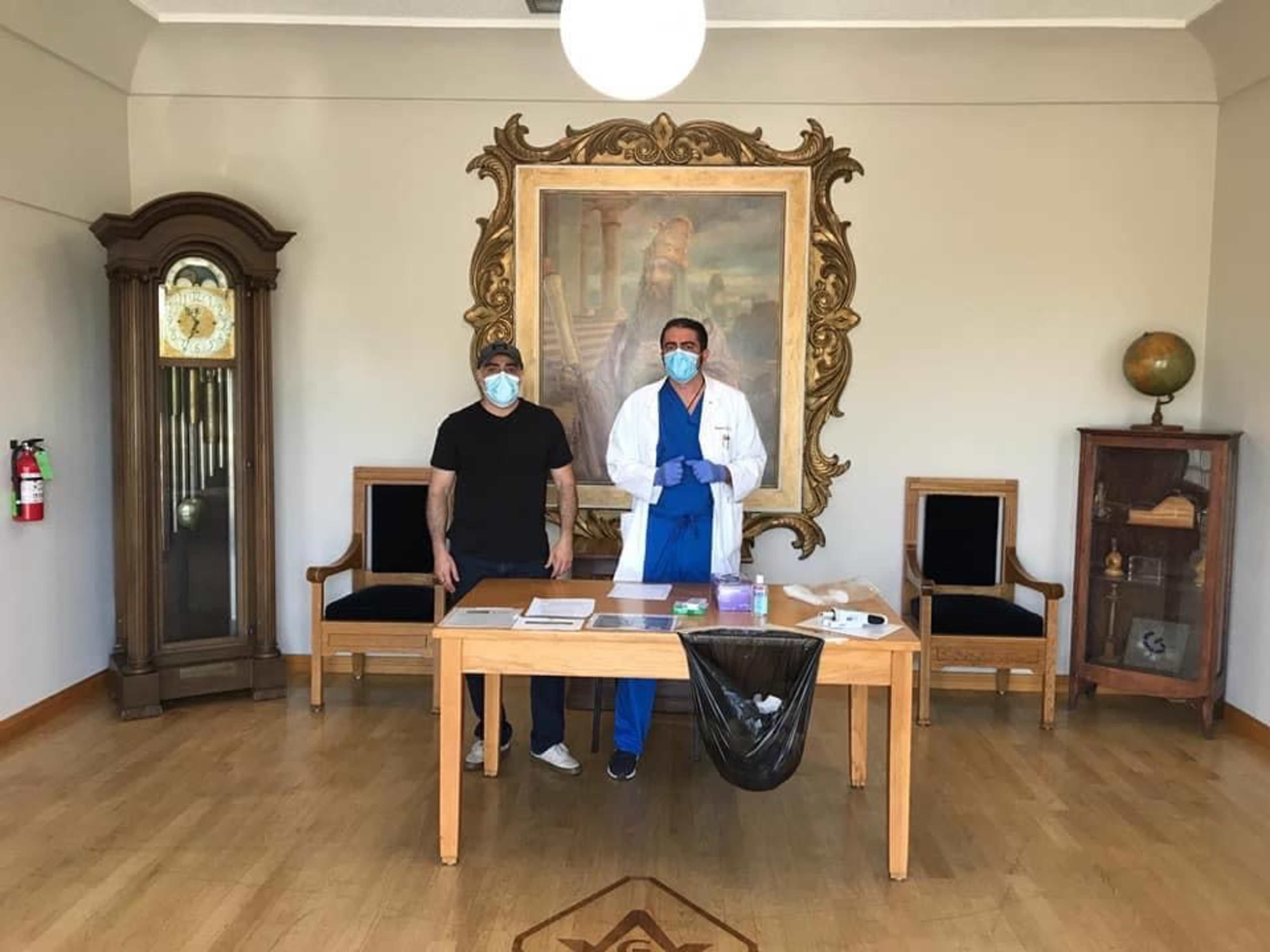 A man in a black shirt stands next to a medical professional in a white coat and blue gloves, both wearing masks, in a room with a large ornate painting and wooden furniture.