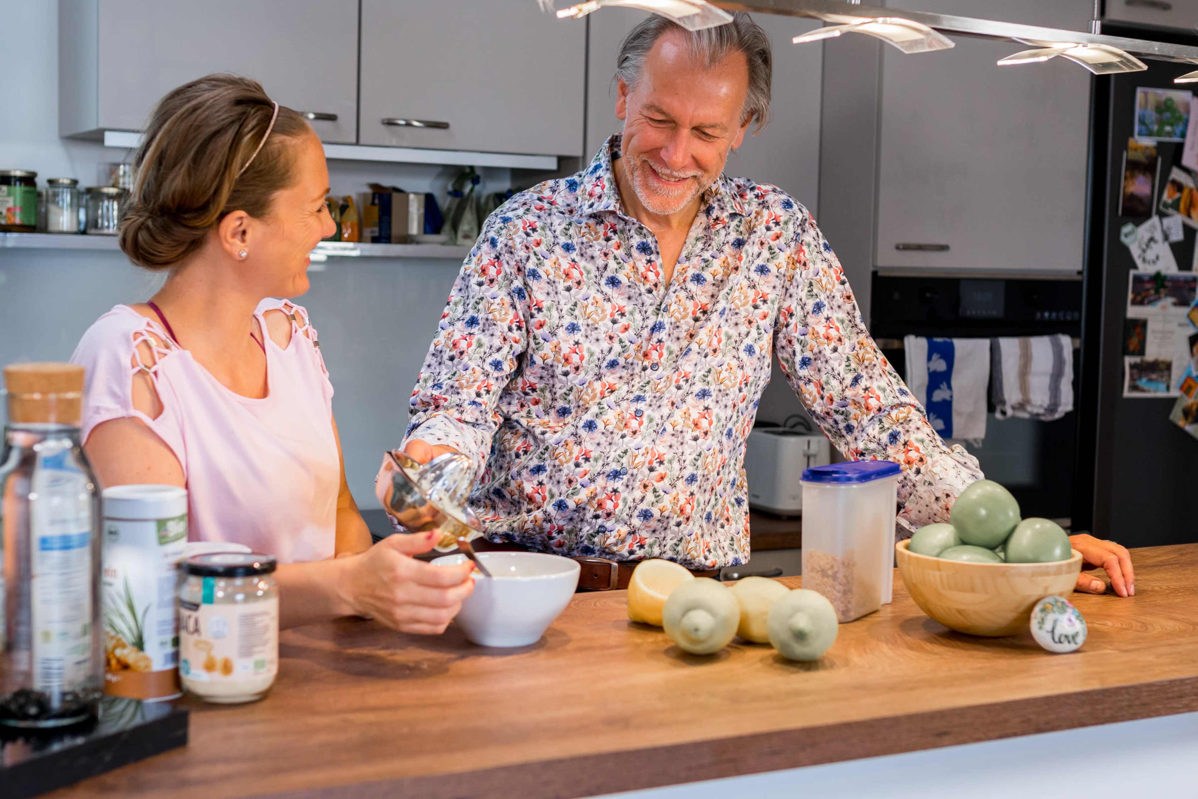 A man and a woman are smiling and interacting in a bright kitchen while preparing food at a wooden countertop filled with ingredients and utensils.