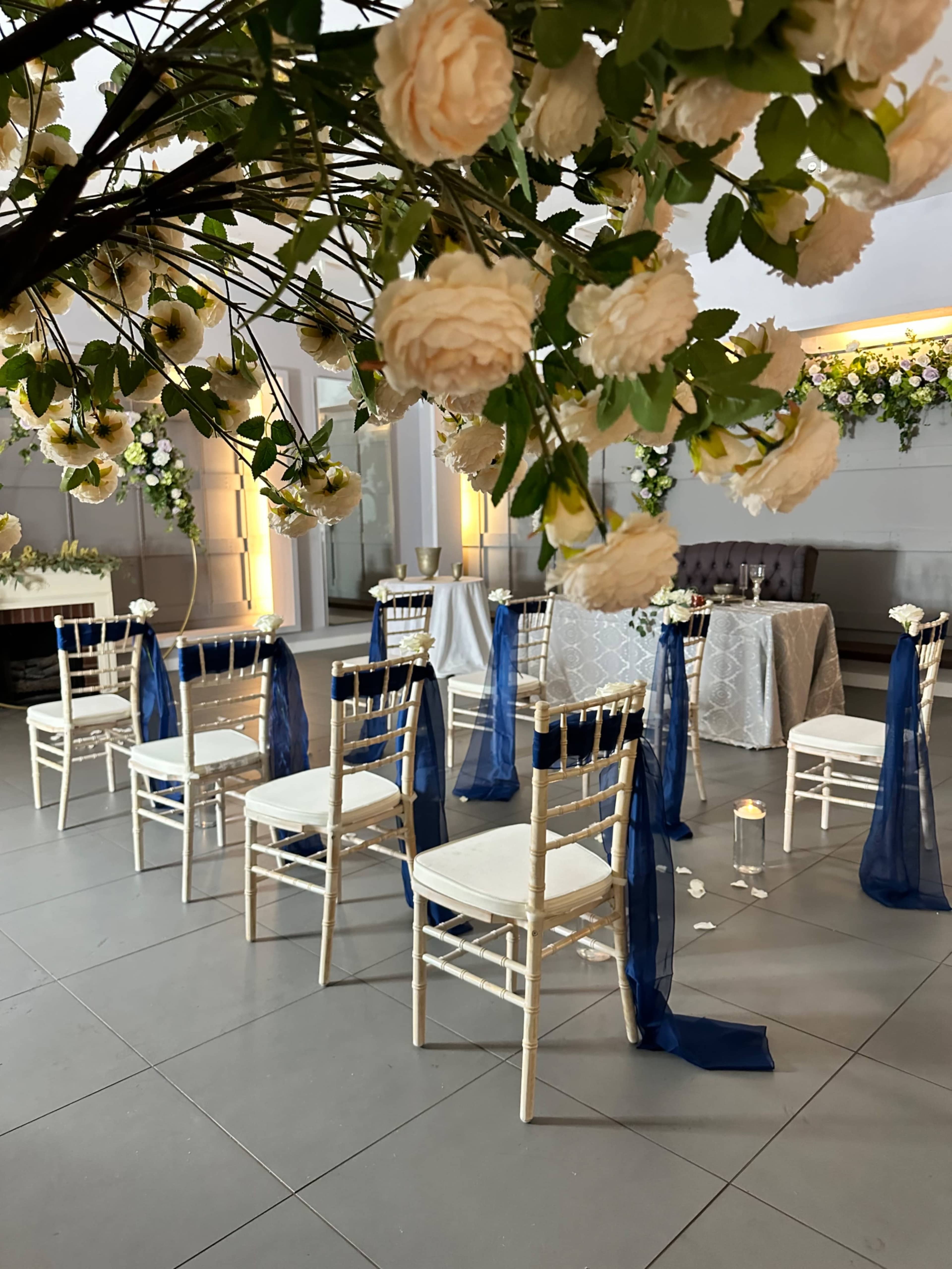 A decorated event space features white chairs arranged with blue sashes beneath an elegant floral arrangement.