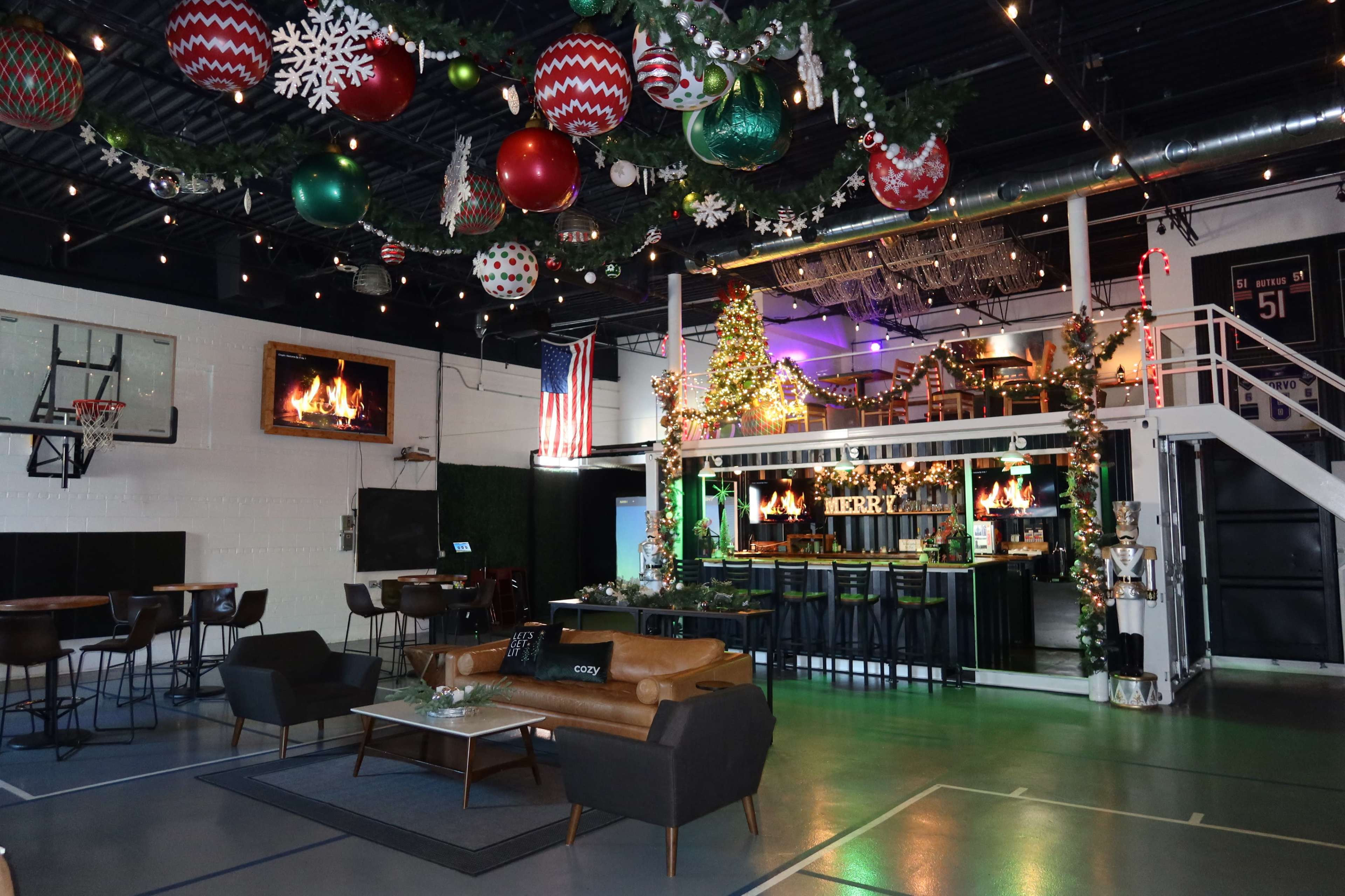 The image shows a festive indoor space decorated with large holiday ornaments, string lights, and a bar area, featuring a basketball hoop in the background.