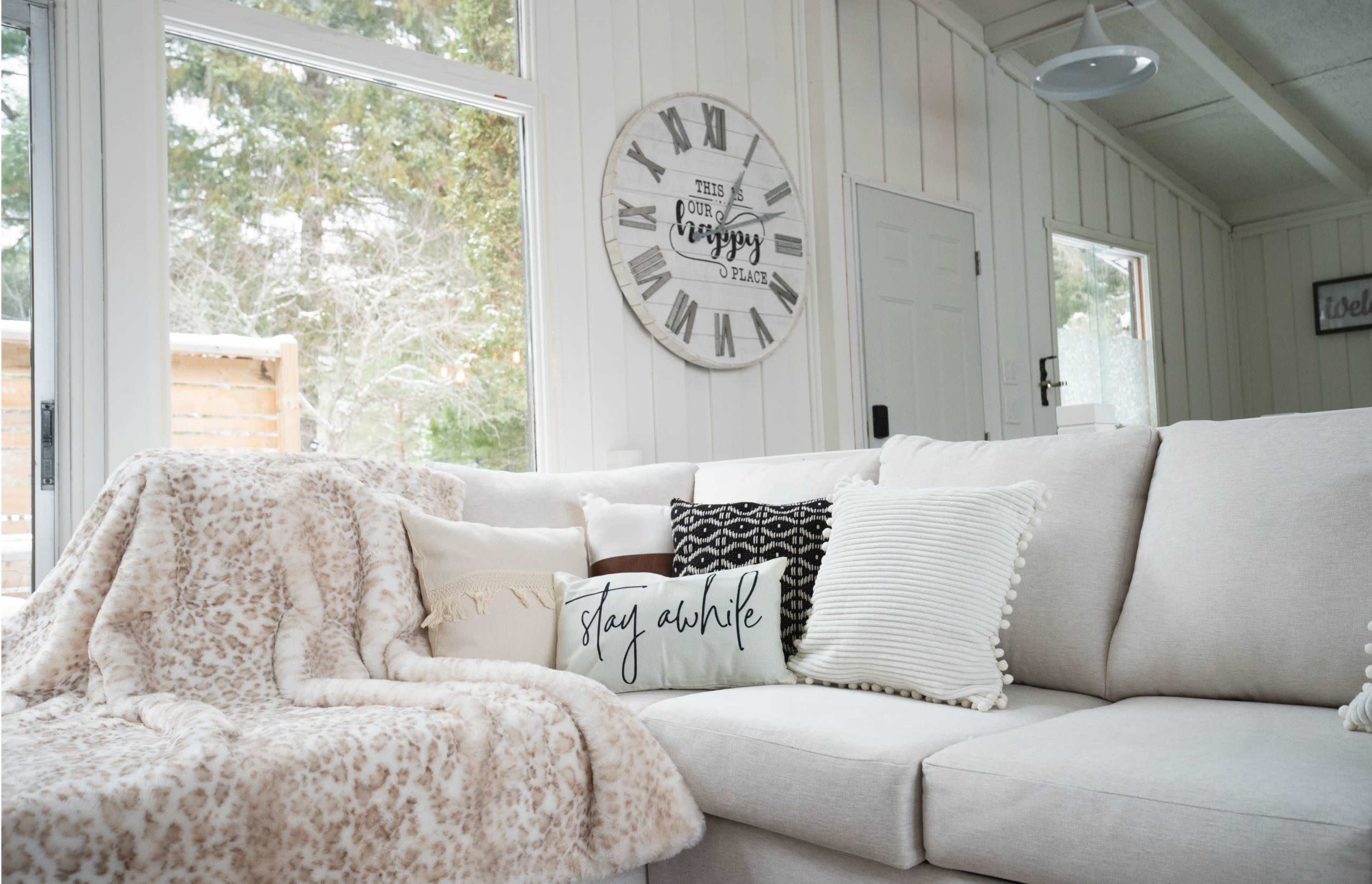 A light-colored sectional sofa is adorned with various decorative pillows and a plush throw blanket, set against a backdrop of large windows and a wall clock.