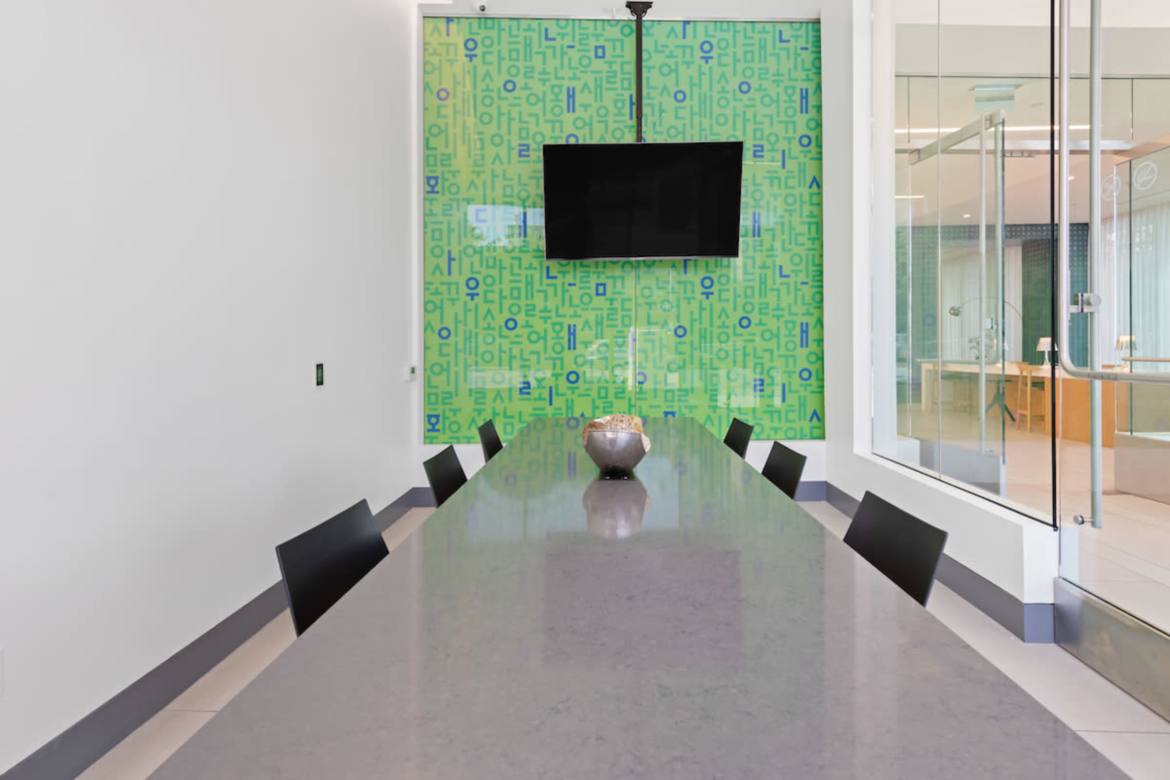 A long conference table with black chairs is positioned in front of a green wall featuring patterned letters and a mounted television.