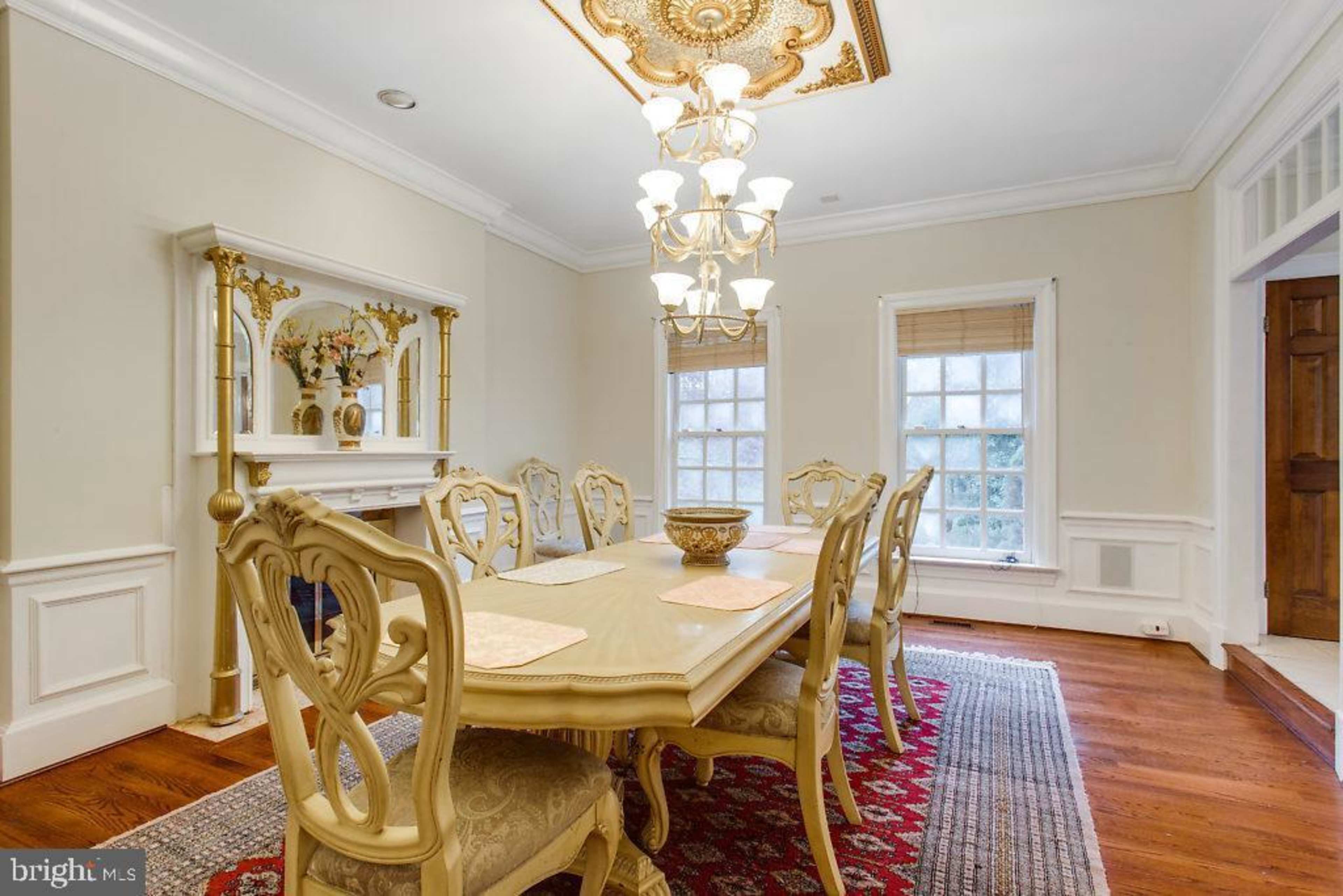 A dining room with a large ornate table, decorative chairs, a chandelier, and windows providing natural light.
